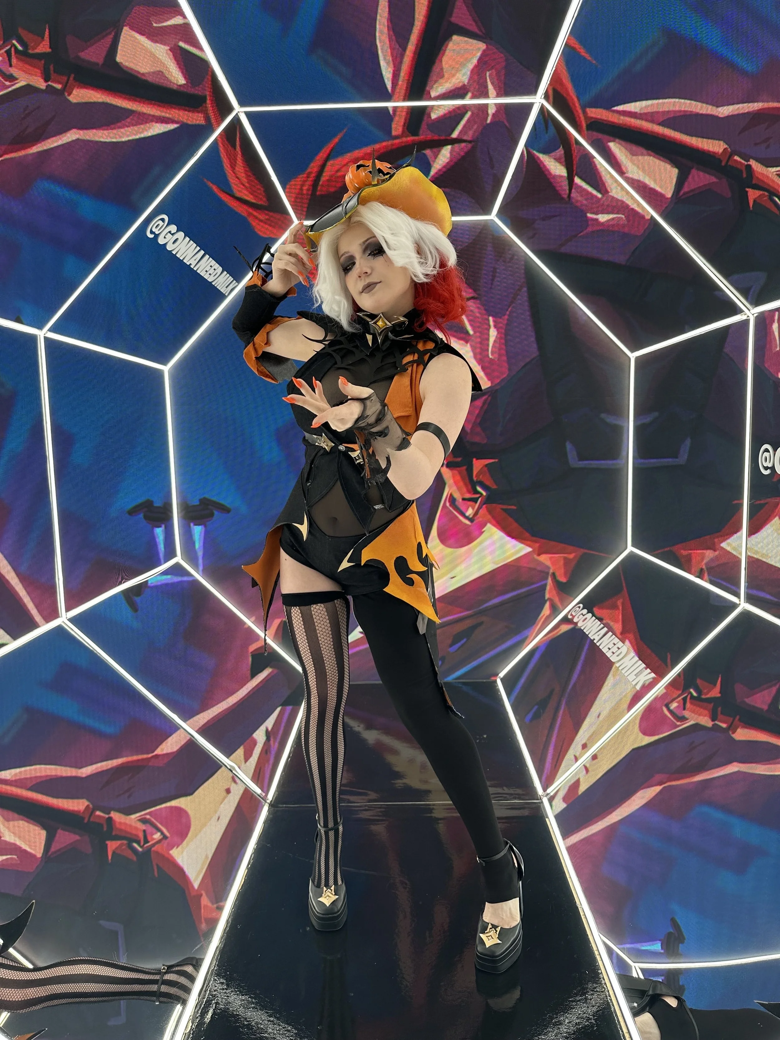 A person dressed in a fantasy costume with black and orange colors, striped thigh-high stockings, and a large yellow hat with black horns, standing in a mirrored room with colorful, abstract digital art on the walls.