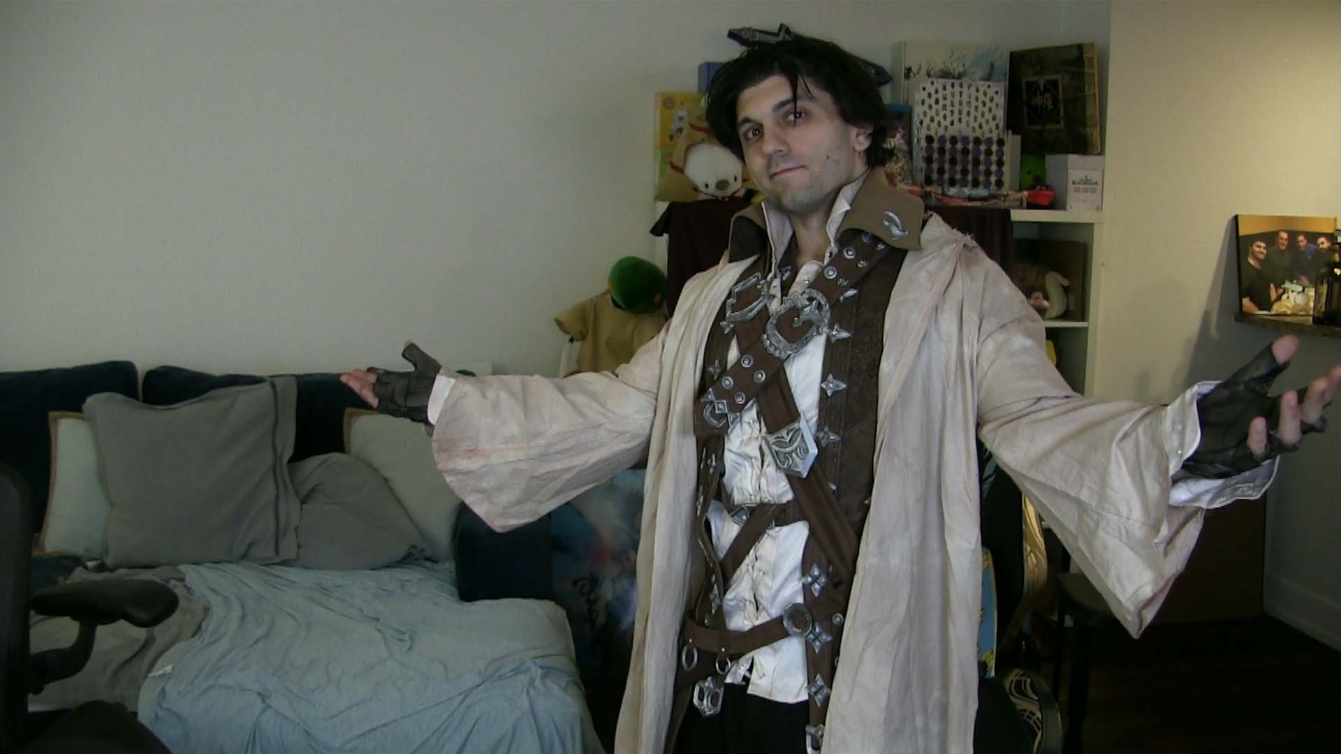 Person dressed as a fantasy character, wearing a tan robe with leather straps and armor, standing in a living room with a beige wall, couch, and shelves with toys and framed photos.