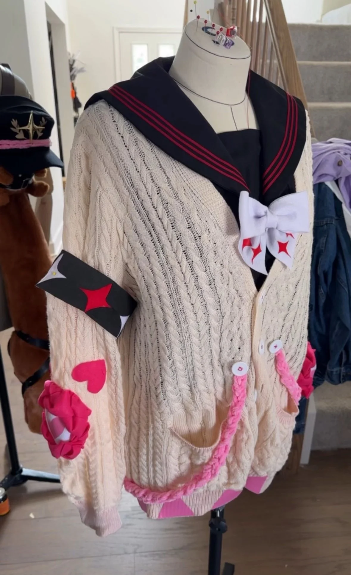 A mannequin dressed in a costume resembling a cosplay outfit, with a knitted cream cardigan featuring pink heart and pink braided string, a black sailor-style collar with red stripes, white bow with red stars, and various patches on the sleeves. In t