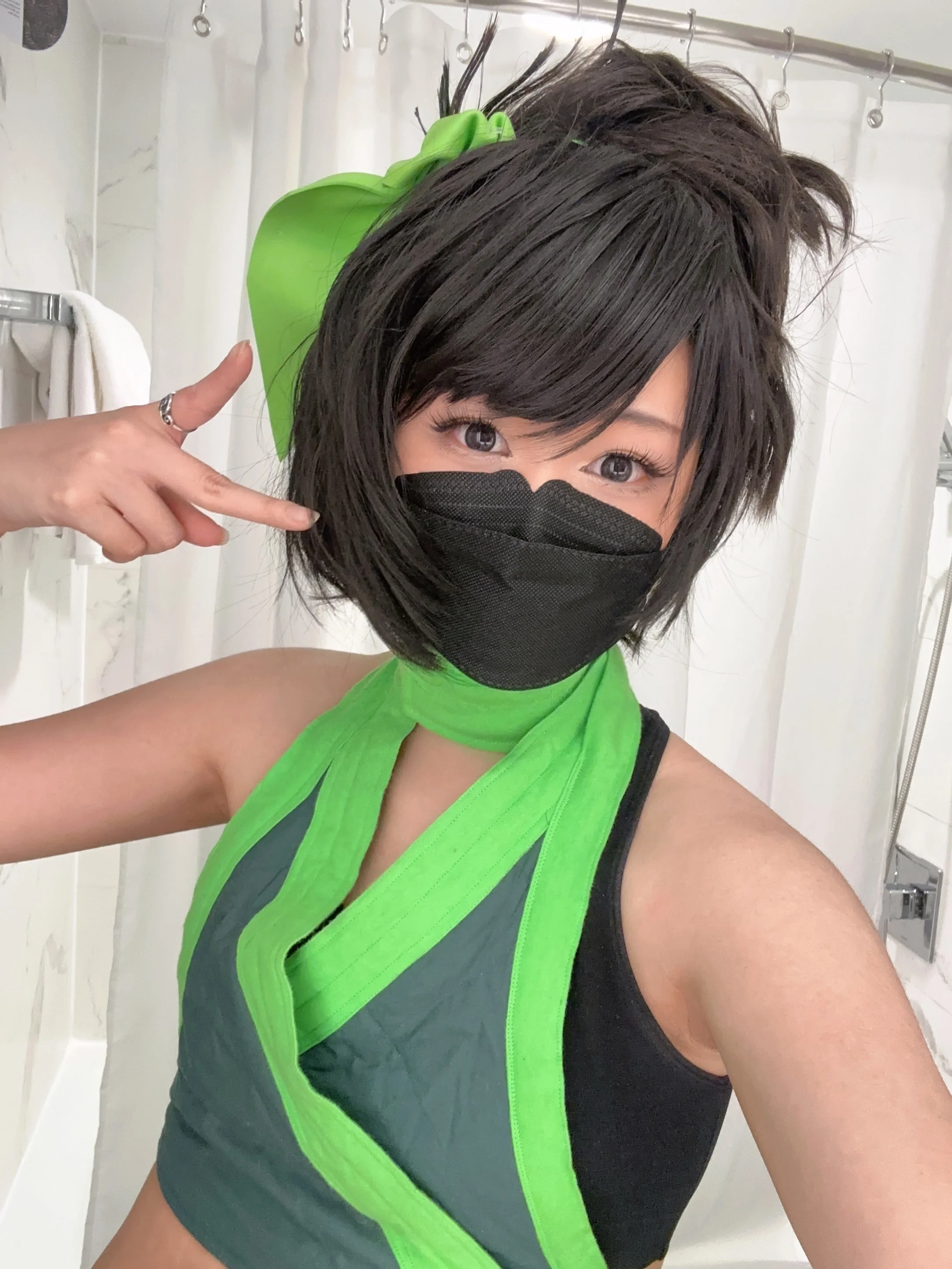 A woman with short dark hair wearing a black face mask, gray and black athletic top with green accents, making a peace sign with her hand, in a bathroom or dressing room.