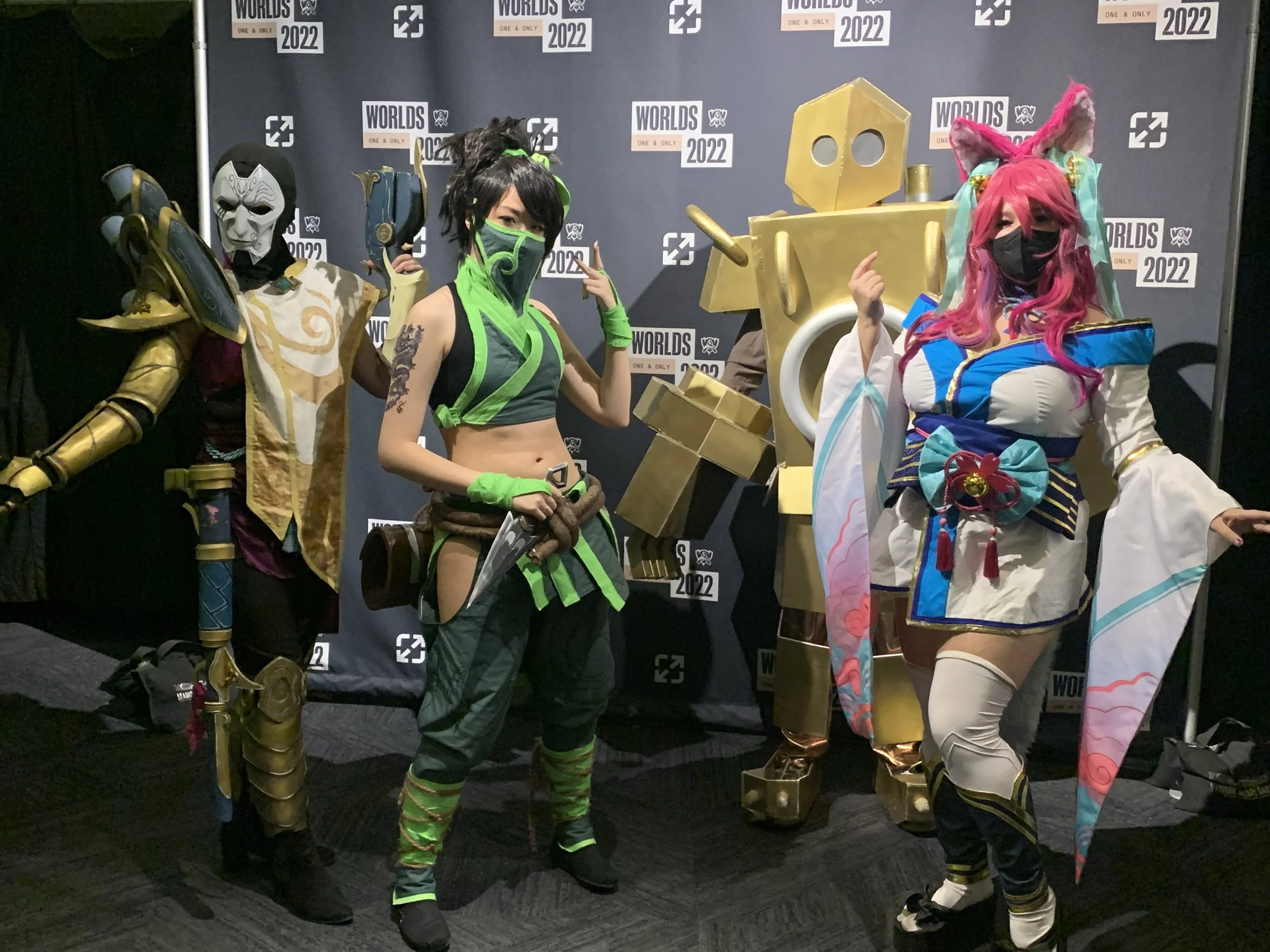 Four people in elaborate costumes at an event with a backdrop displaying 'WORLDS 2022'. The first person on the left is dressed as a warrior with gold armor and a mask. The second person is in a green and black ninja-style outfit with a face mask and