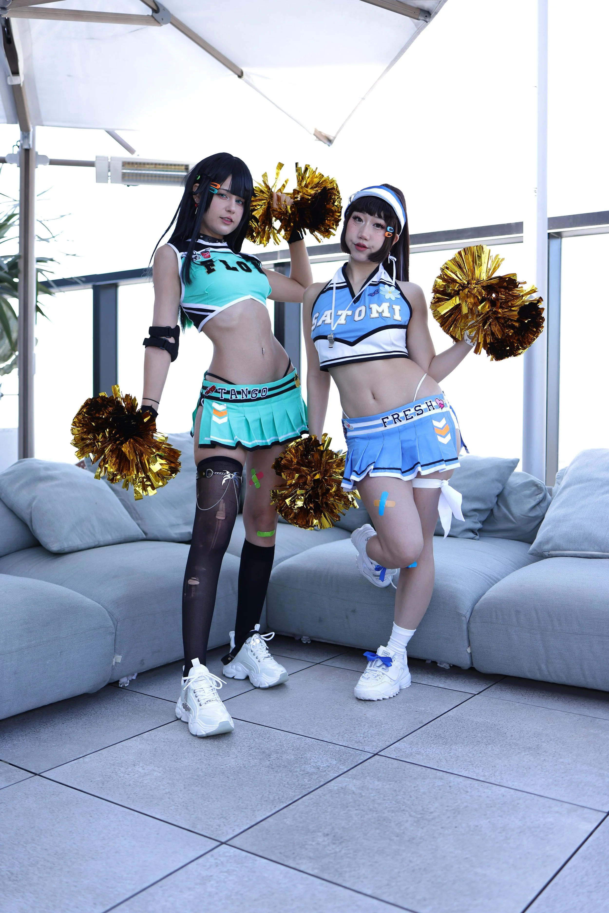 Two women dressed in cheerleading outfits with metallic pom-poms, standing on a tiled balcony with a gray sofa behind them.