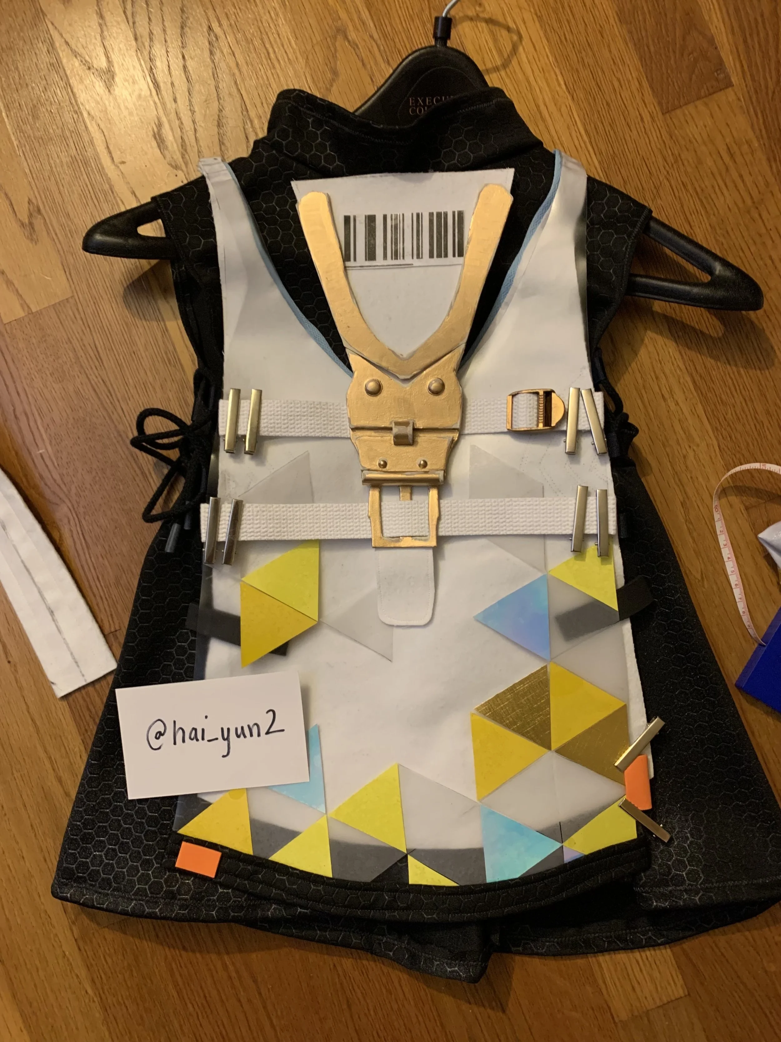 A white harness with yellow, blue, and gold geometric shapes attached, placed on a black sports bag on a hardwood floor.