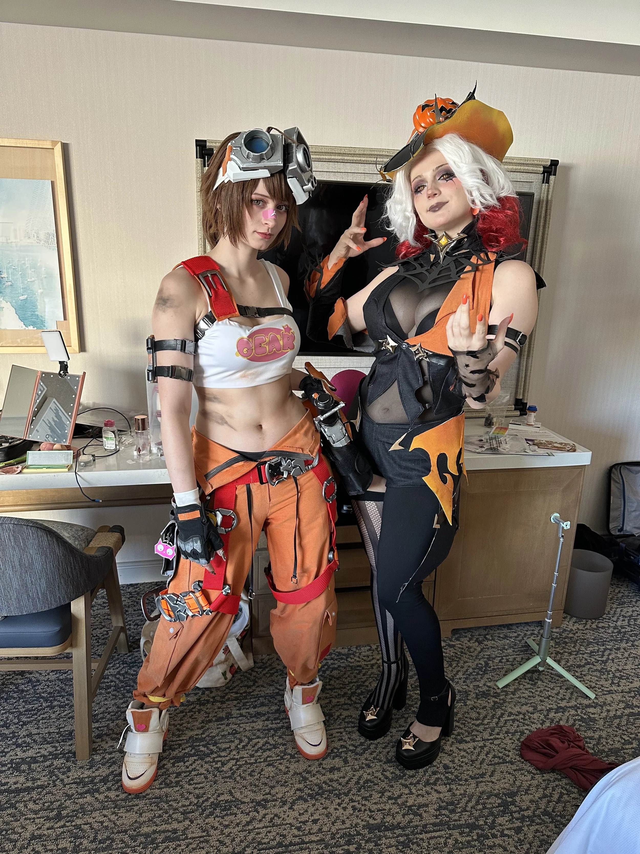 Two women in elaborate costumes posing in a hotel room, with one dressed as Overwatch character Tracer and the other as Halloween witch.