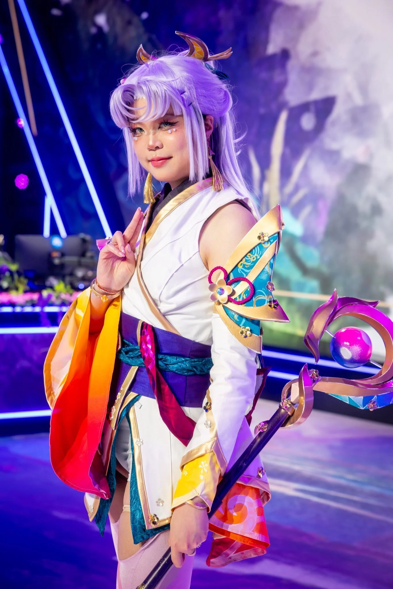 A person dressed in a colorful fantasy costume with lavender hair, golden horns, and a detailed staff, standing in a vibrant, futuristic setting.
