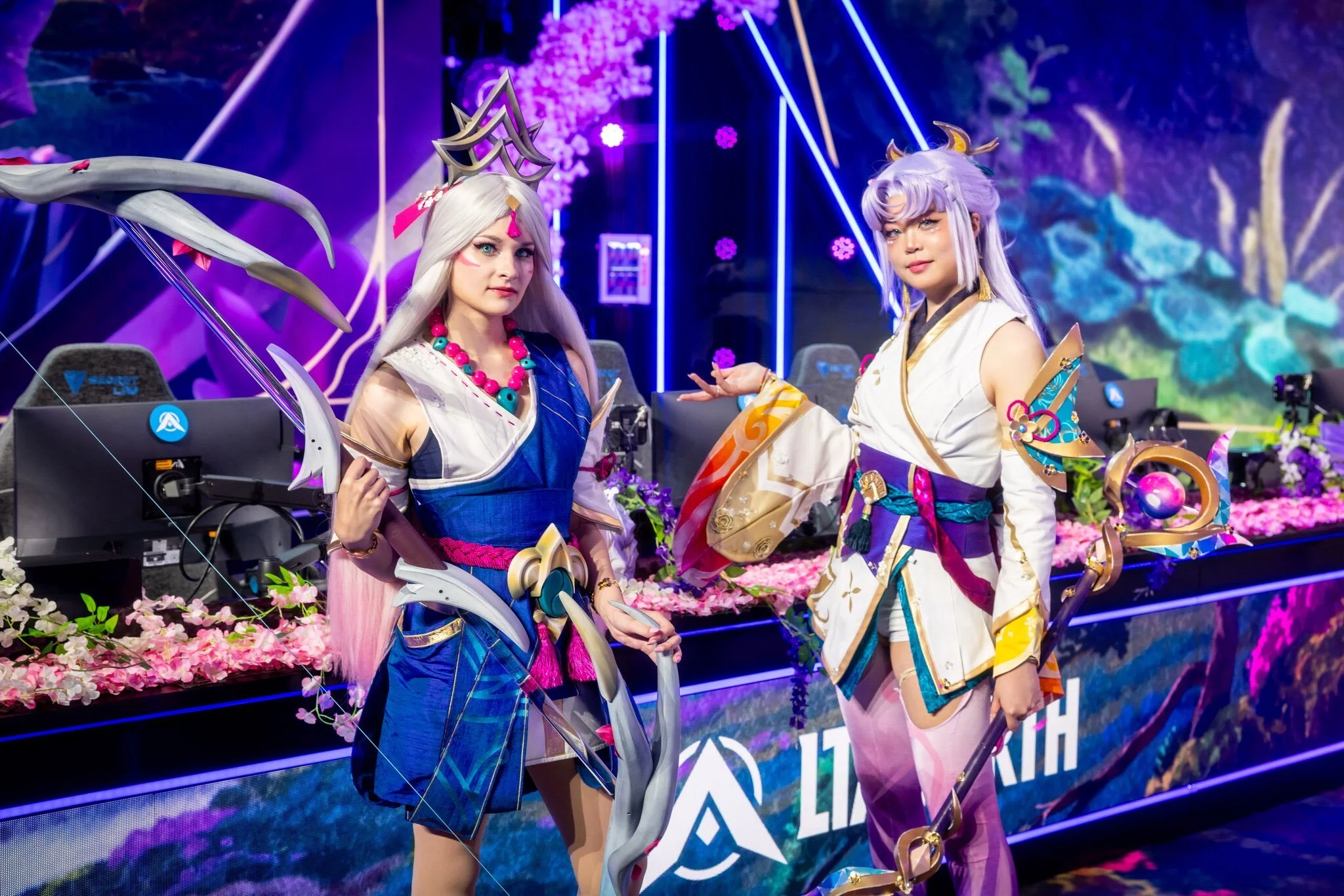 Two cosplayers dressed as fantasy characters pose in front of a colorful background at an event, with a floral and illuminated setting.