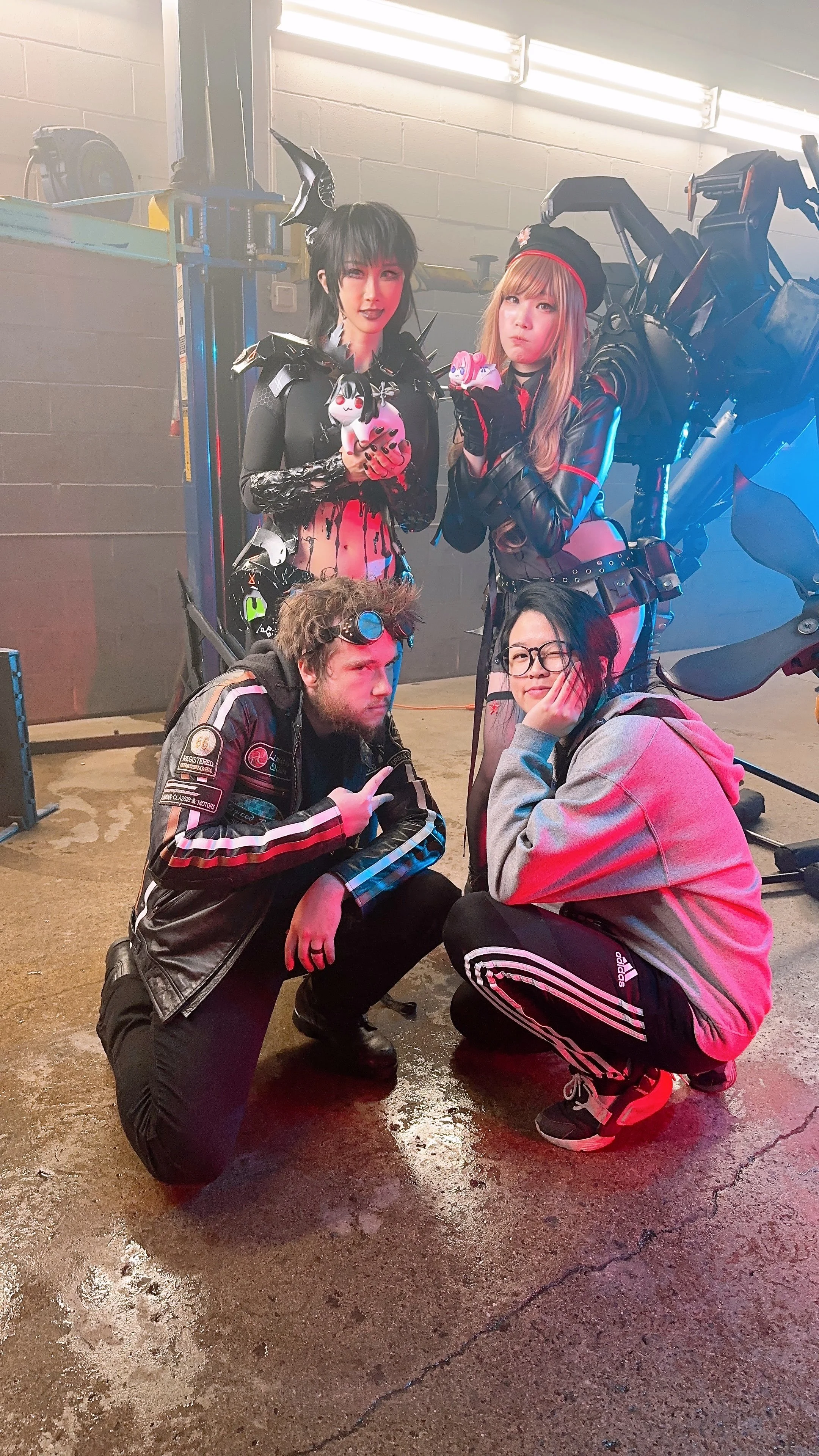 Four people in strange costumes in a dimly lit garage, two women standing in the back holding plush toys, one man crouching in front making a peace sign, and one woman squatting with glasses, all posing for the photo.