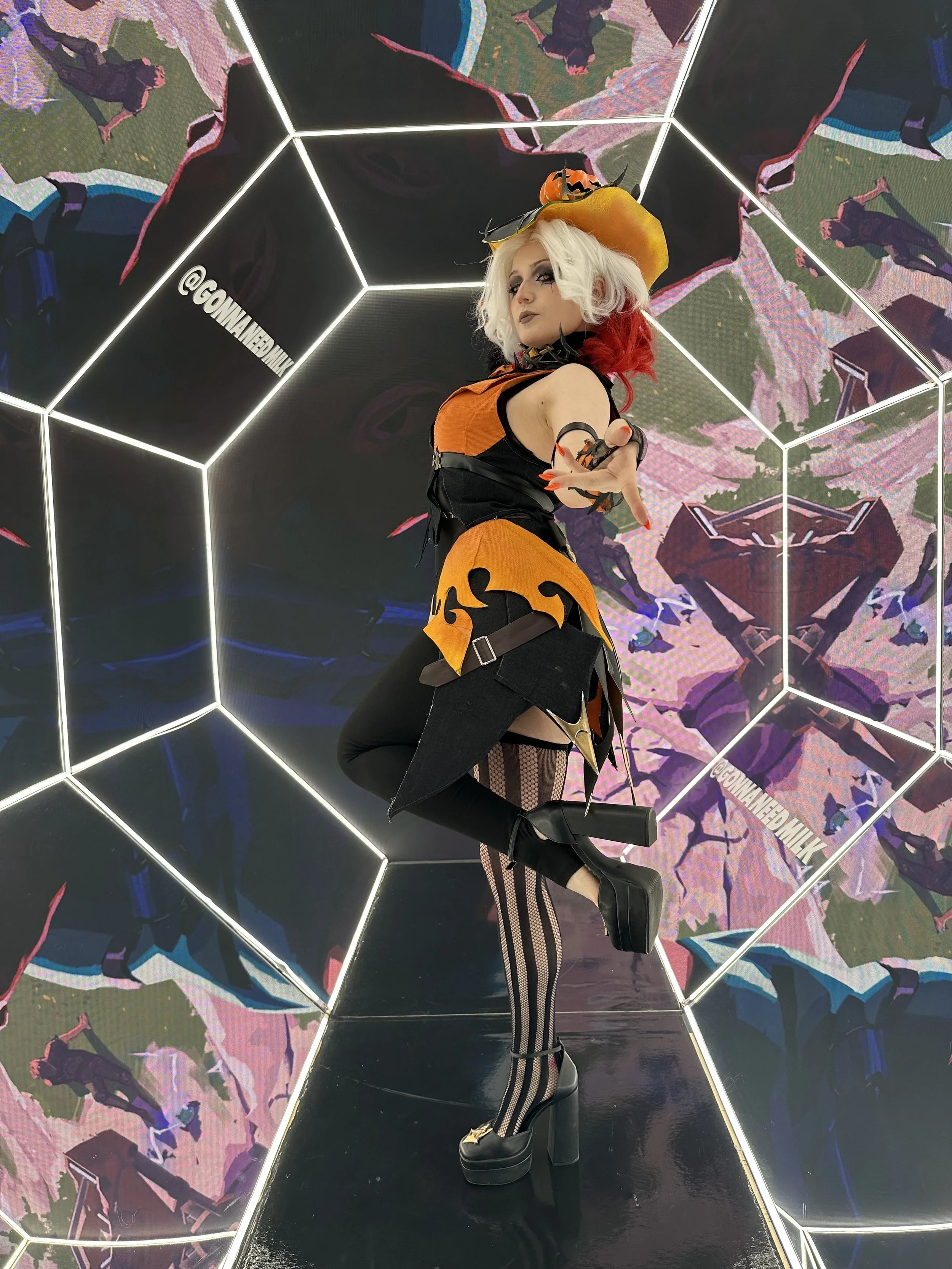 A person dressed in a Halloween-inspired costume with a witch hat, orange, black, and red hair, and black makeup, posing in a mirrored room with bright white light outlines and digital artwork on the walls.
