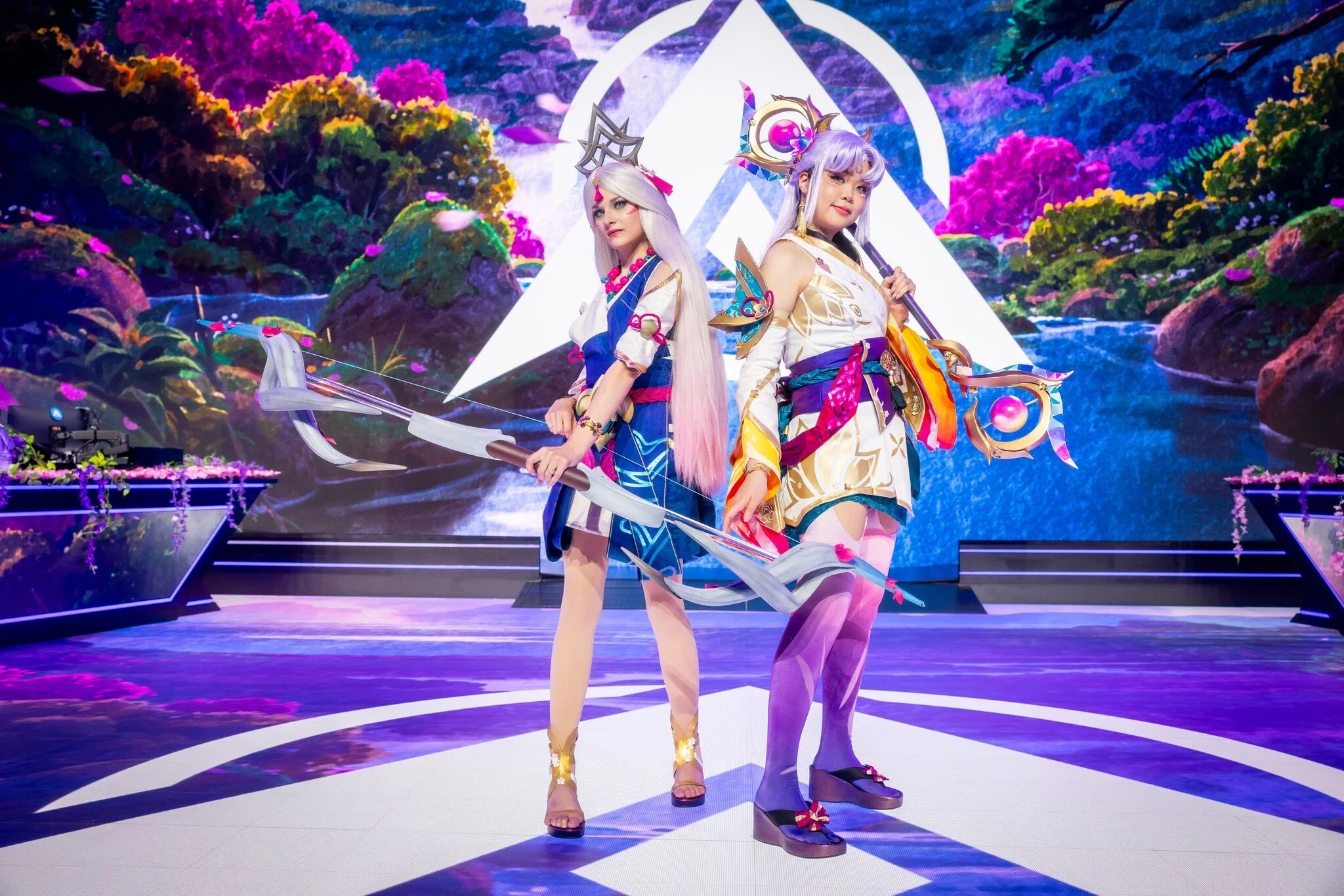 Two women in colorful, fantasy-themed costumes standing on a stage with a vibrant, animated backdrop of a fantasy landscape featuring trees, water, and a large glowing symbol.