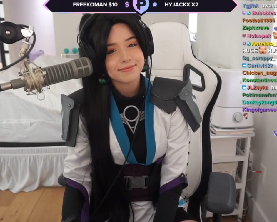 A young woman dressed as a character from Overwatch, wearing a white and black suit with blue accents and shoulder armor, sitting in front of a microphone during a live streaming session.