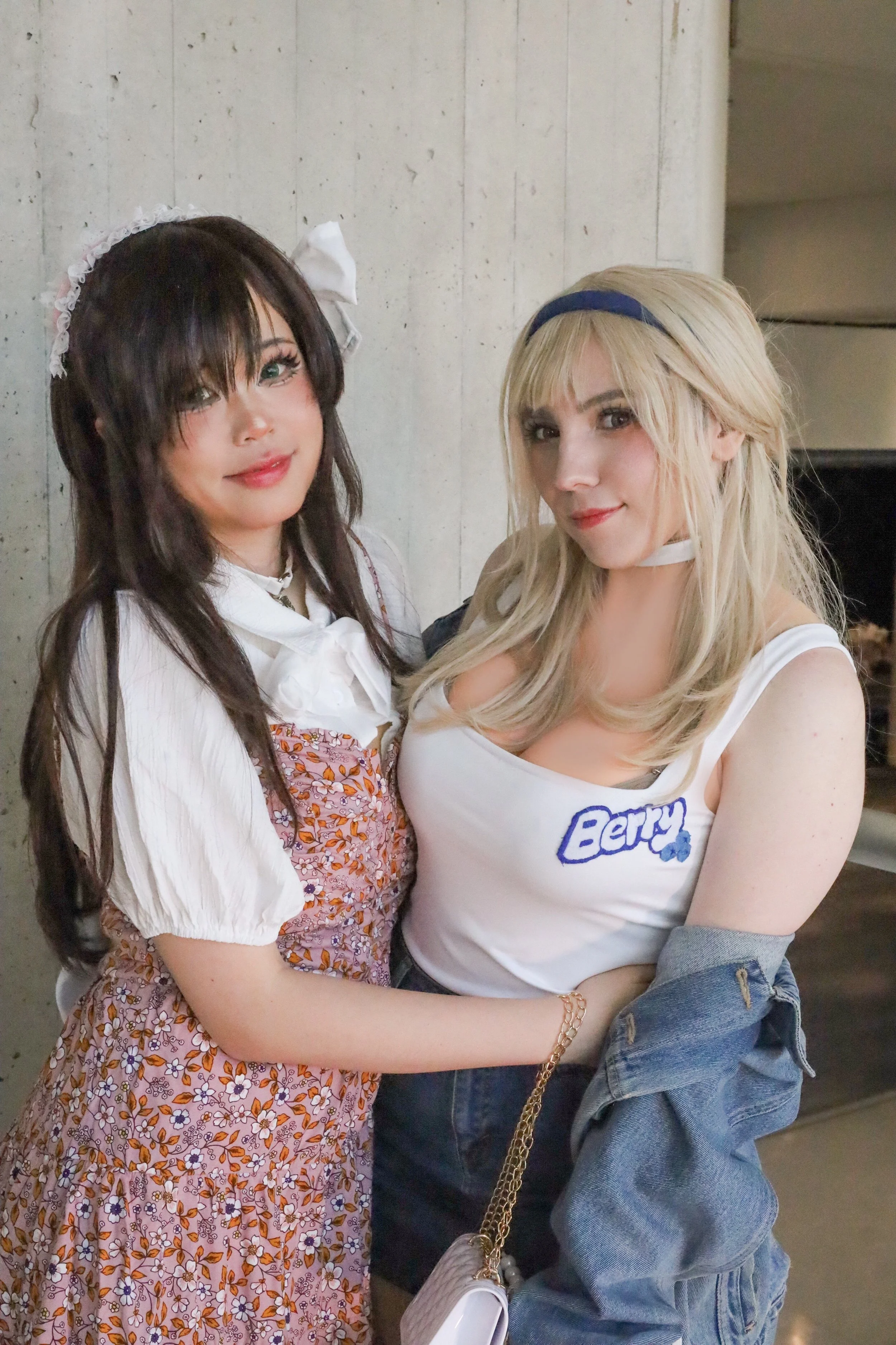 Two women standing close together, one with dark brown hair and a white and pink floral dress, and the other with blonde hair, wearing a white tank top with 'BERRY' printed on it and a denim jacket draped over her arm.