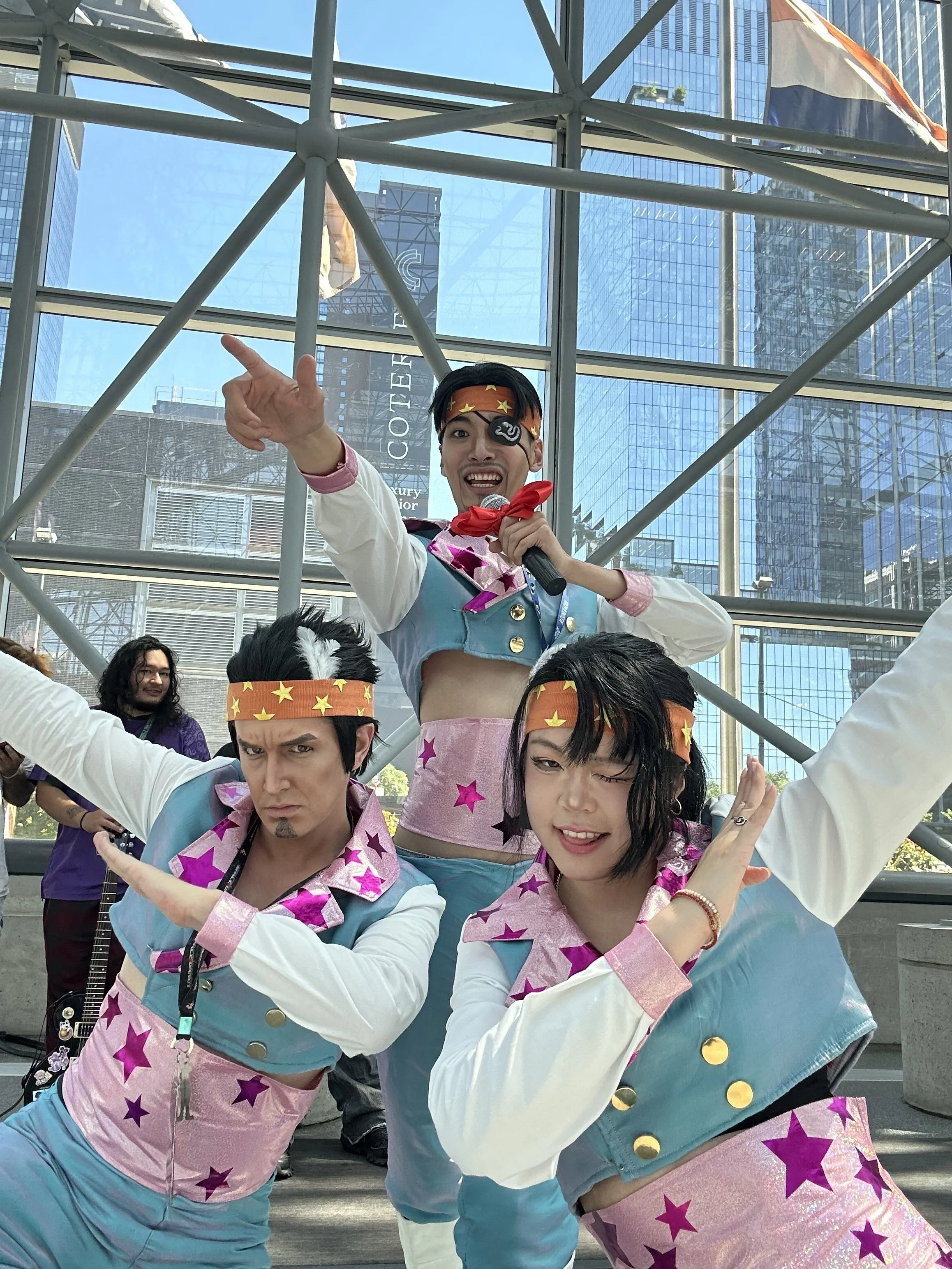 Three cosplayers dressed in colorful, star-themed costumes striking dynamic poses inside a glass building with tall skyscrapers in the background.