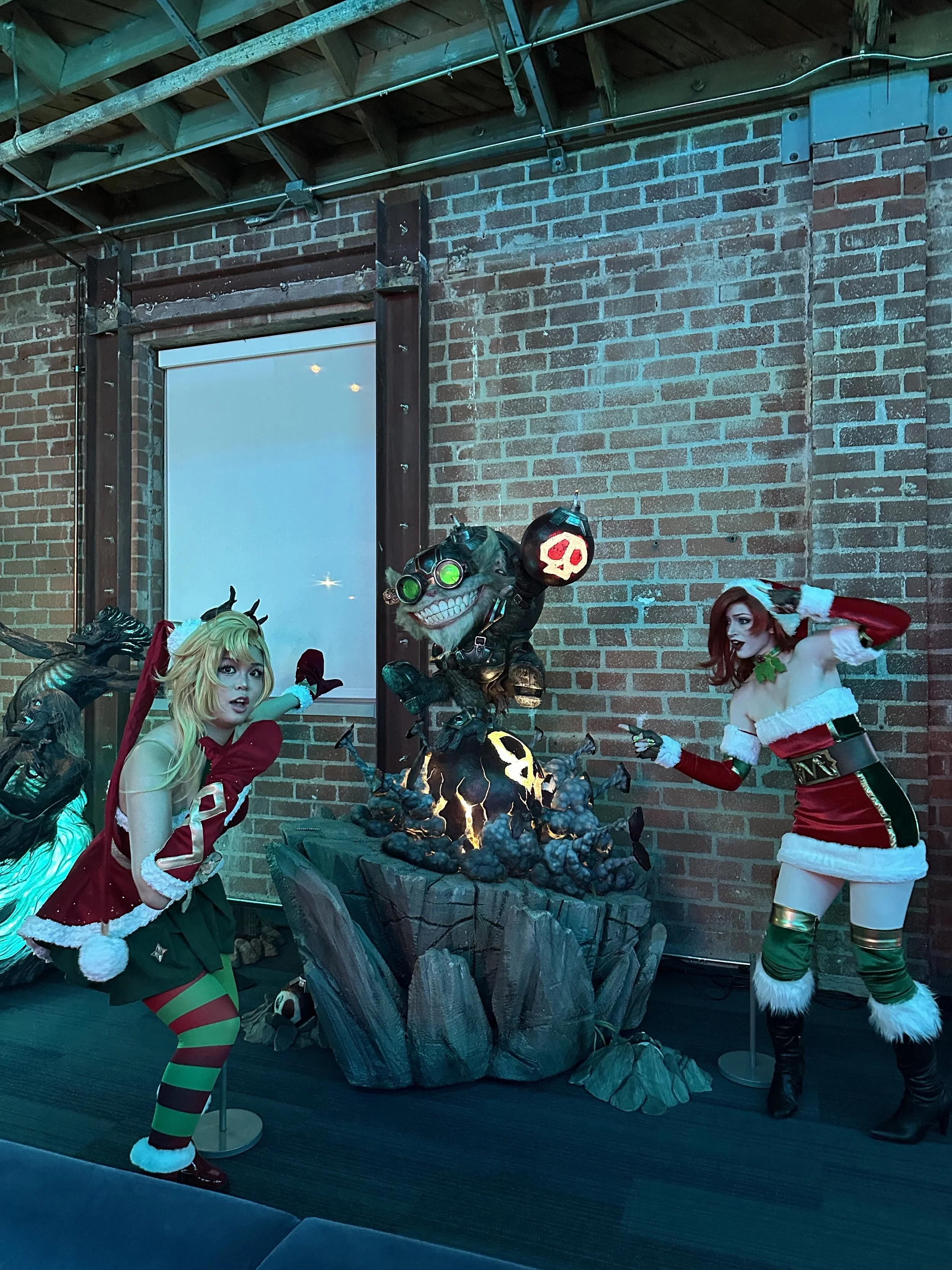 Two women in Christmas-themed costumes pose around a large, animated Halloween decoration of a spooky creature emerging from a rocky base, inside a brick-walled room.