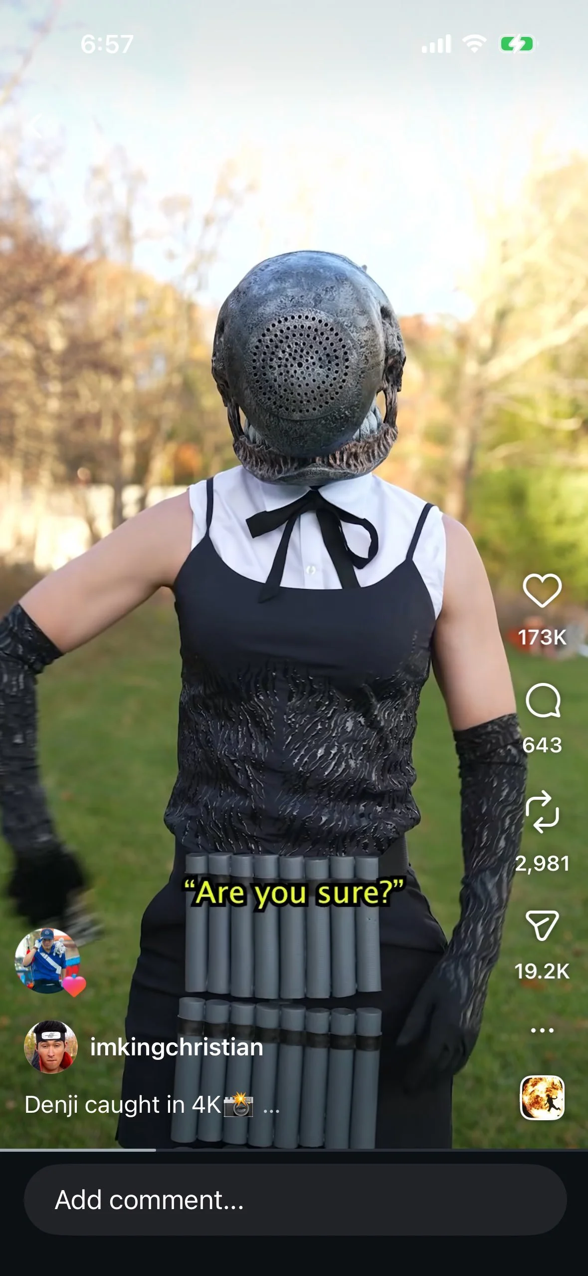 Person wearing a black mask resembling a helmet, a black and white dress with a bow, and red glowing items on waist, standing outdoors with trees in the background.