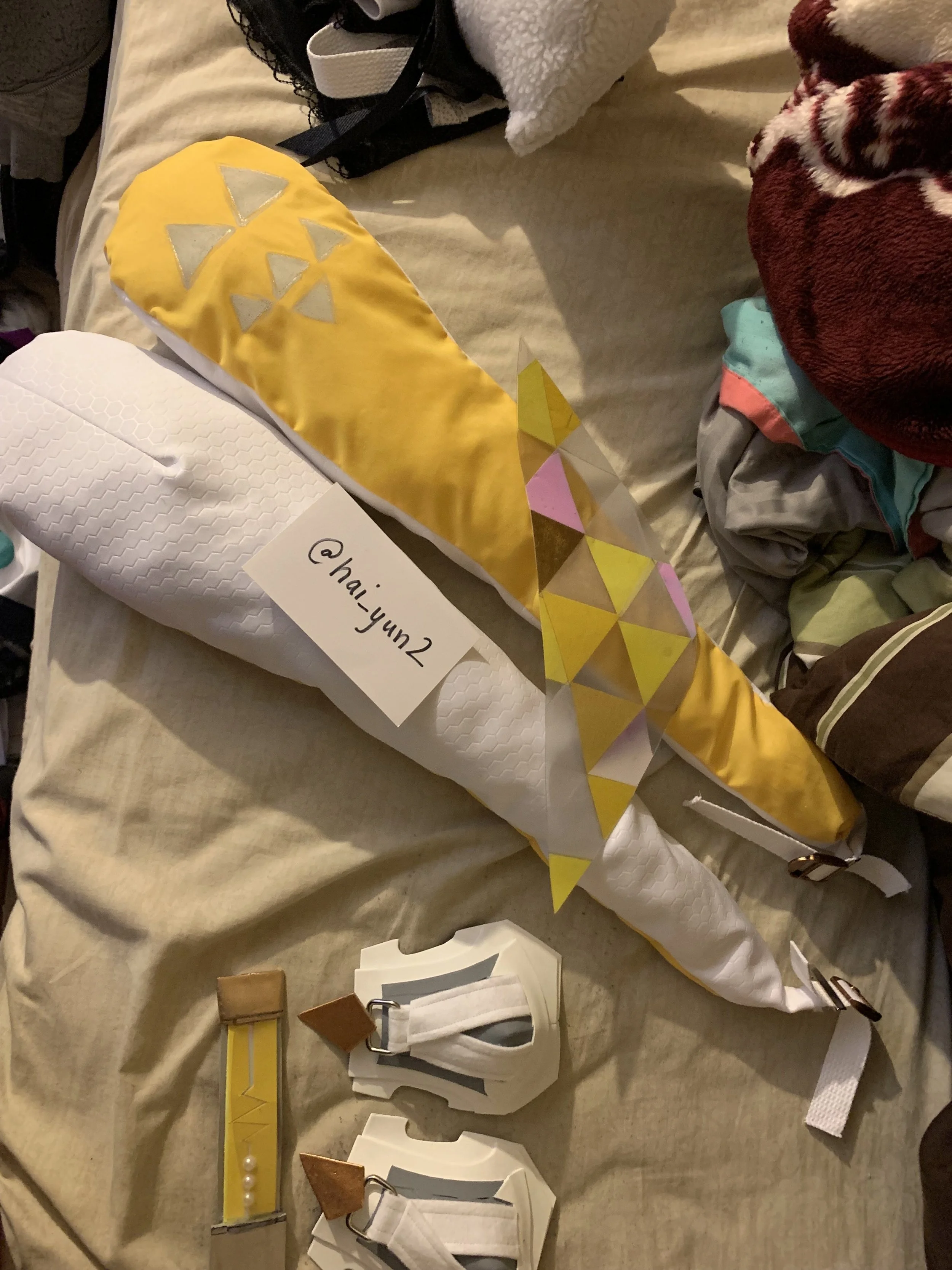 Decorative long paper cone resembling a rocket, with yellow, gold, pink, and clear triangle patterns. Next to it are white name tags with a buckle and string, and a yellow ribbon with white pearls attached. The background shows a bed with clothes and
