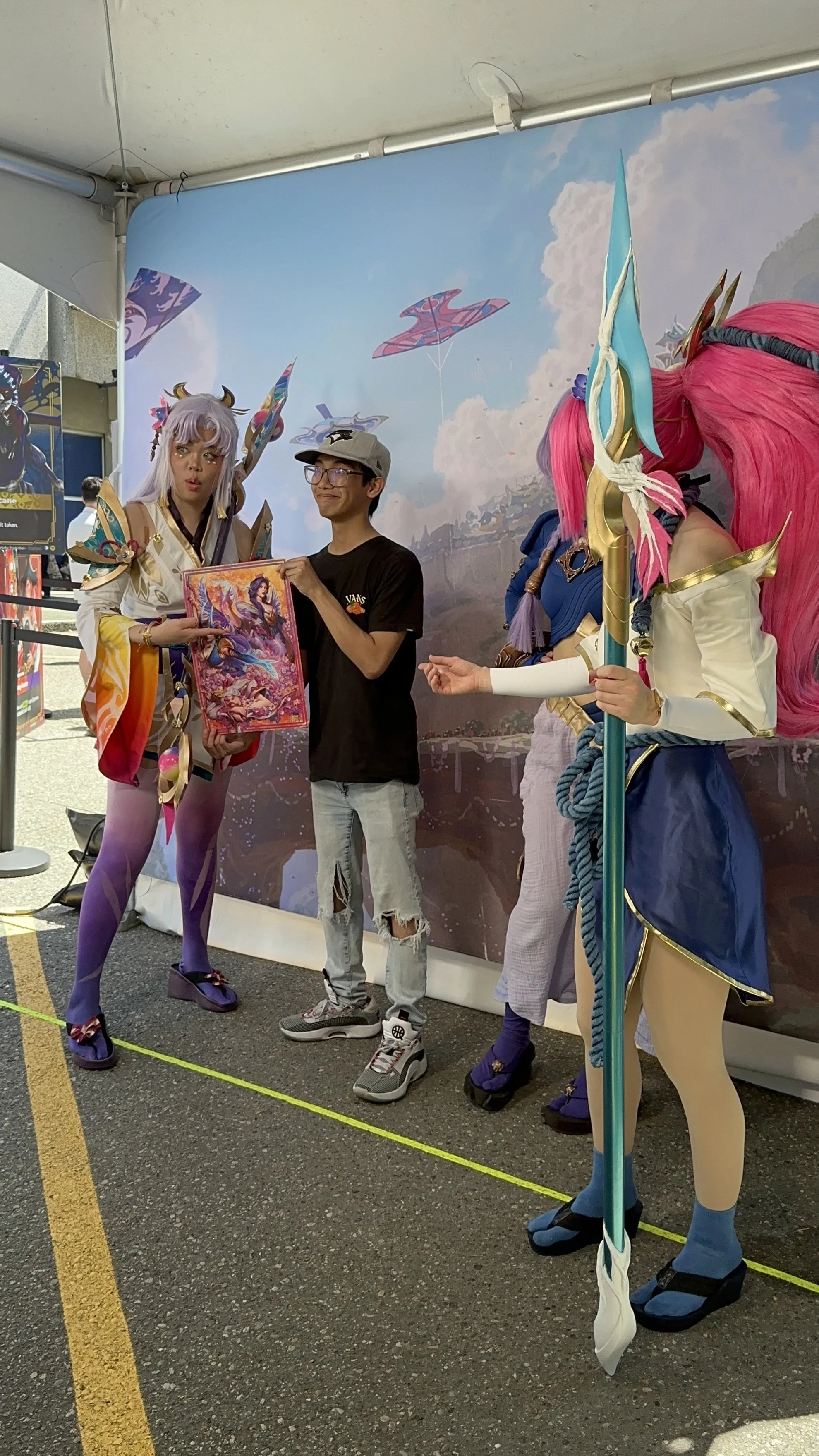 A person is posing with two cosplayers dressed as fantasy characters at an event, with a colorful fantasy-themed backdrop and kites flying in the sky.