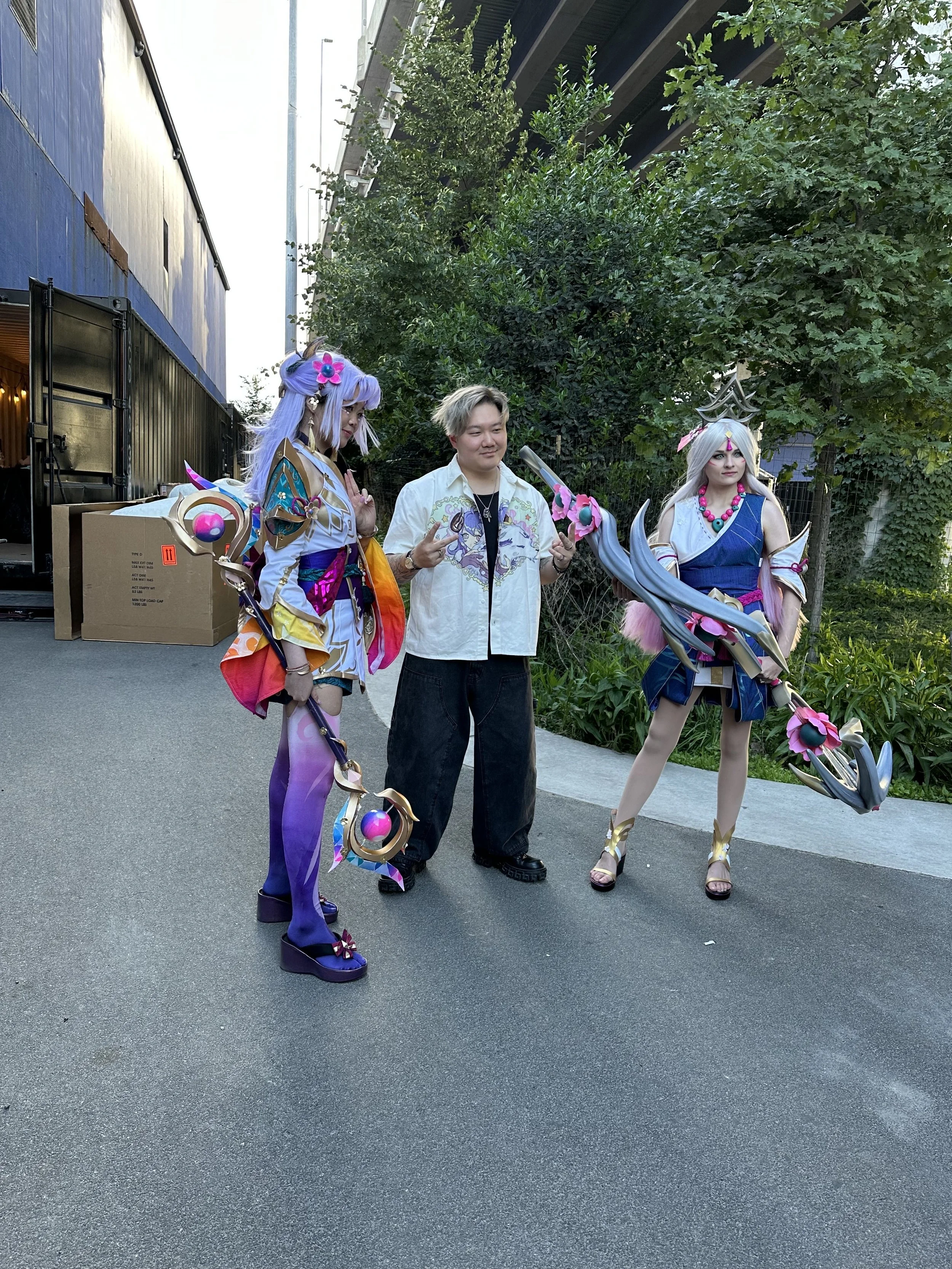 Two cosplayers dressed as fantasy characters pose with a man making peace signs outdoors next to greenery and a building.