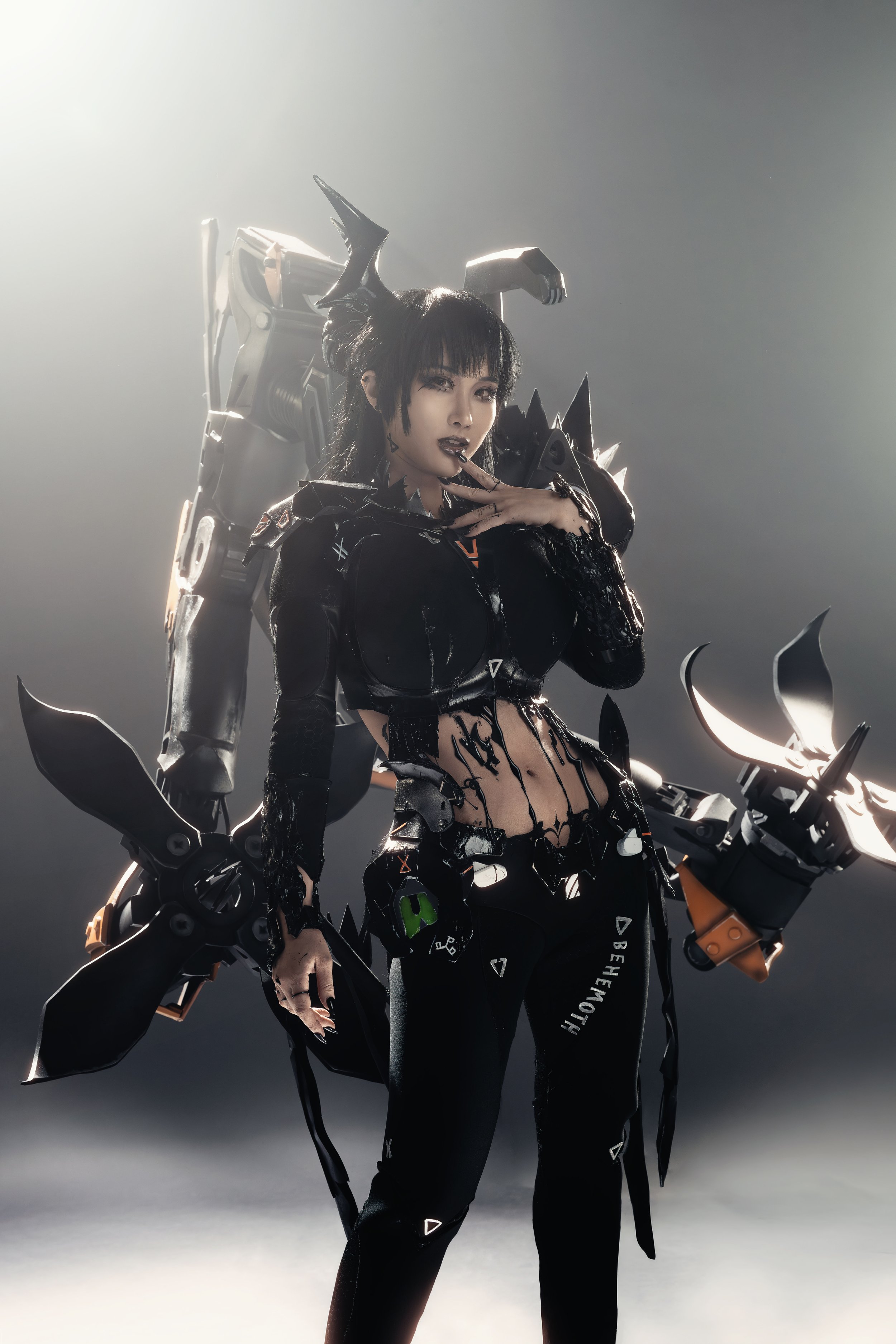 A woman in a black, futuristic costume with mechanical armor and weaponry, striking a pose with her hand near her face.