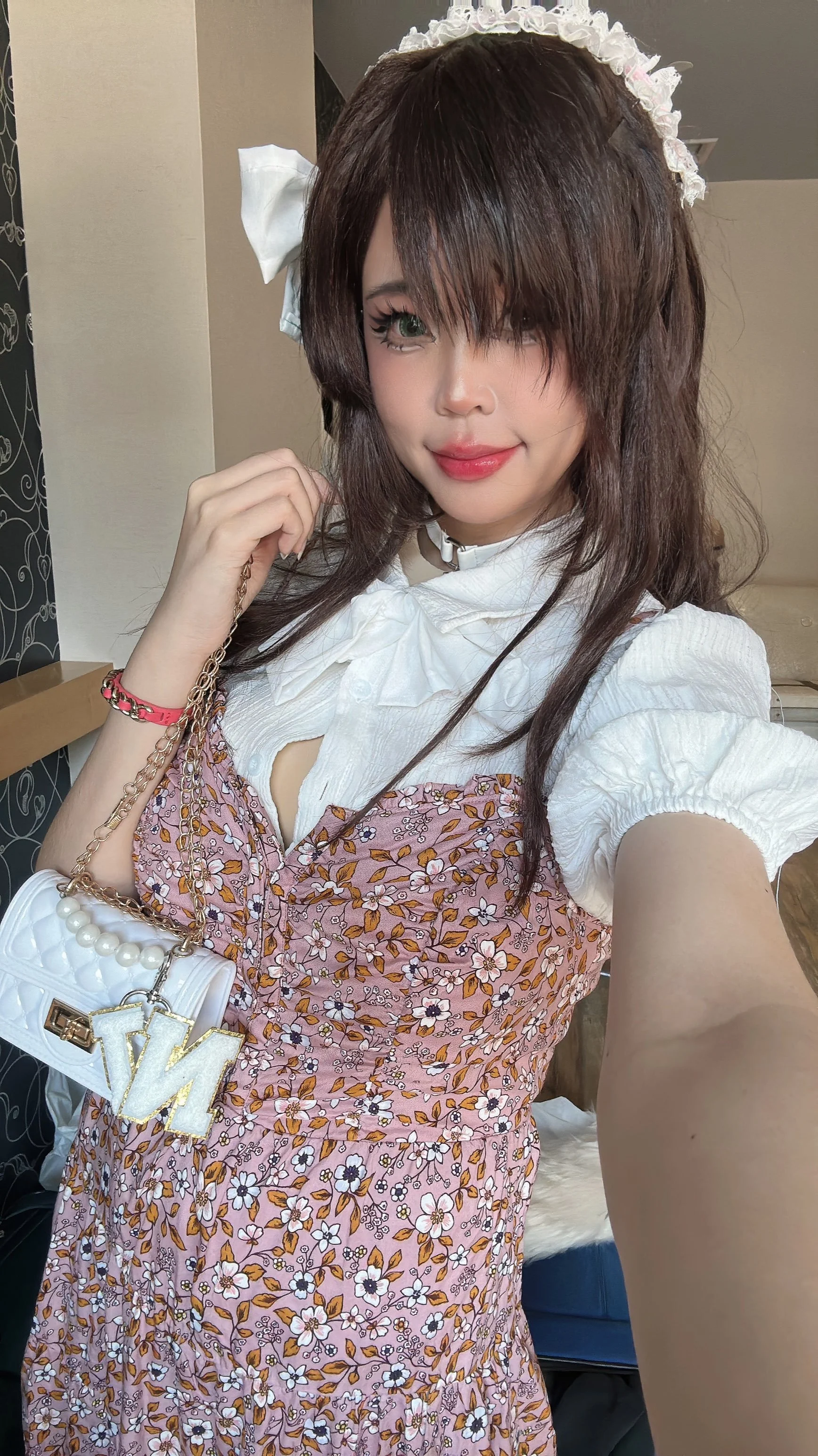 A young woman taking a selfie, dressed in a white blouse with puffed sleeves layered under a pink floral dress, wearing a white headband with lace, holding a white purse with pearl and chain details, and standing indoors.
