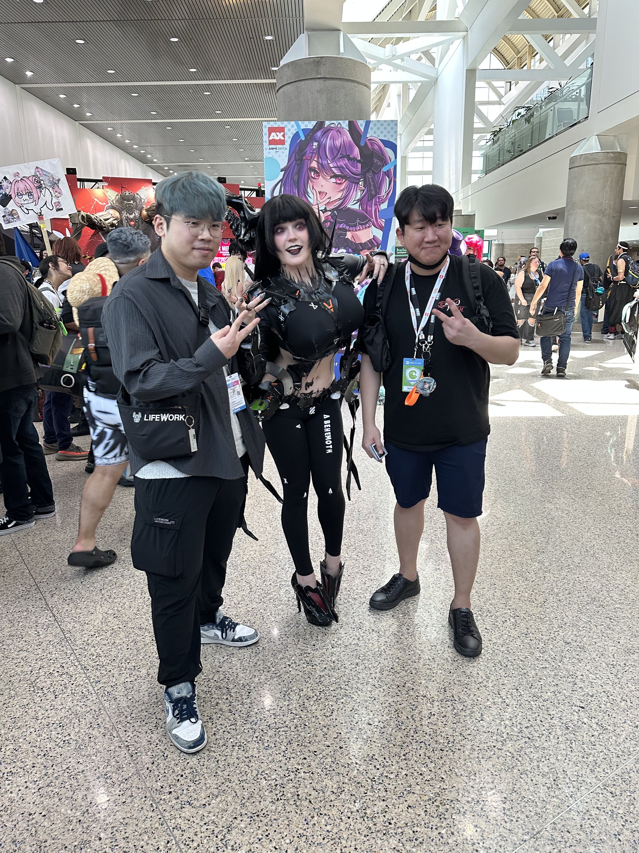 Three people posing at an anime convention, two young men and a woman dressed in a black cyberpunk costume, standing in front of colorful anime posters and crowded convention hall