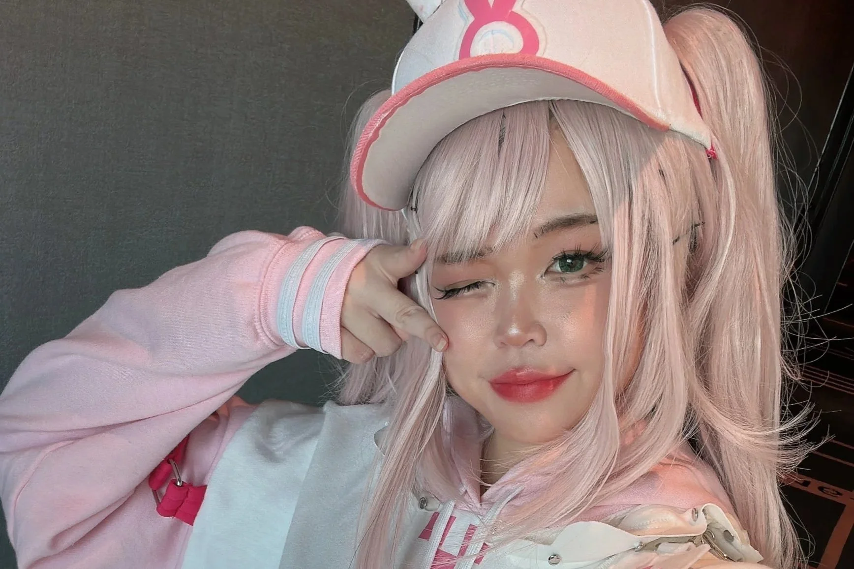 A young woman with pink hair in pigtails, wearing a pink and white cap and a pink hoodie, making a peace sign with her fingers near her eye and smiling.