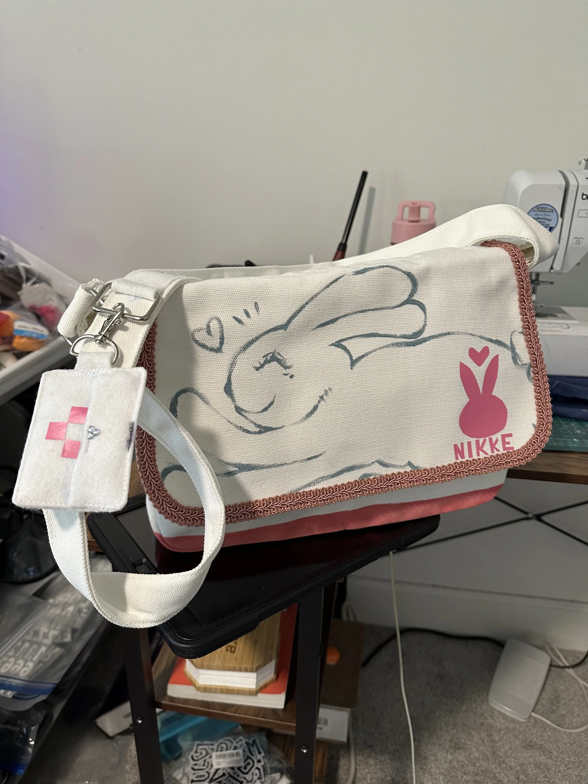 A Bag with a drawing of a happy bunny rabbit and the word NIKKE, placed on a small black table in a room with sewing and crafting supplies.