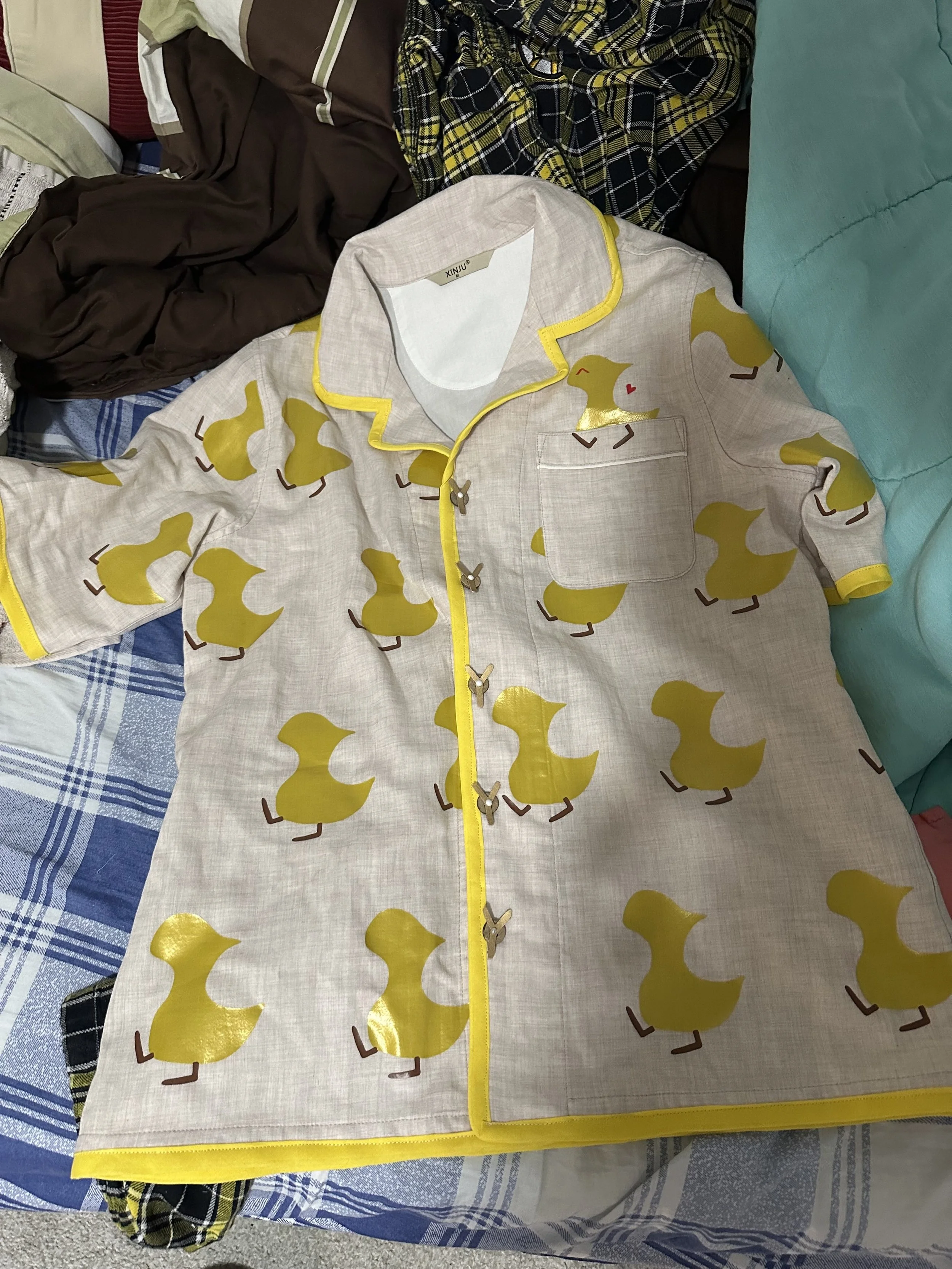 A cream-colored pajama top with yellow duck pattern, yellow trim, and a front pocket, laid on a cluttered surface with other clothes.