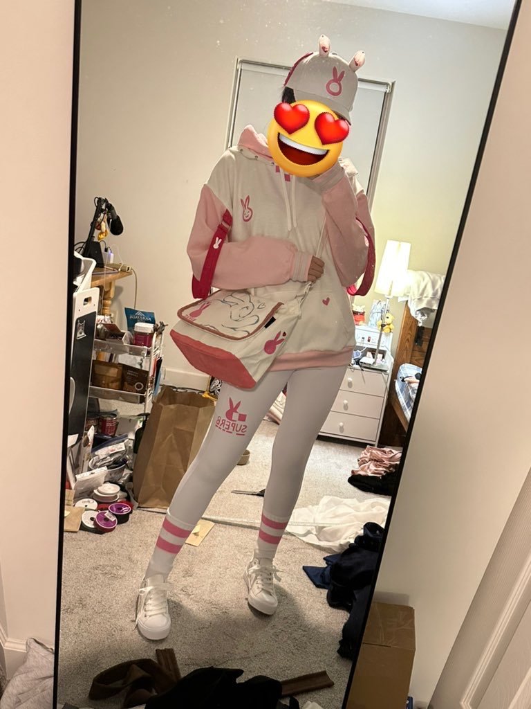 Person taking a mirror selfie in a bedroom, wearing a pink and white hoodie, matching leggings, and a hat with bunny ears. The face is covered with a heart-eyes emoji. The room is messy with clothes, books, and other items scattered around.