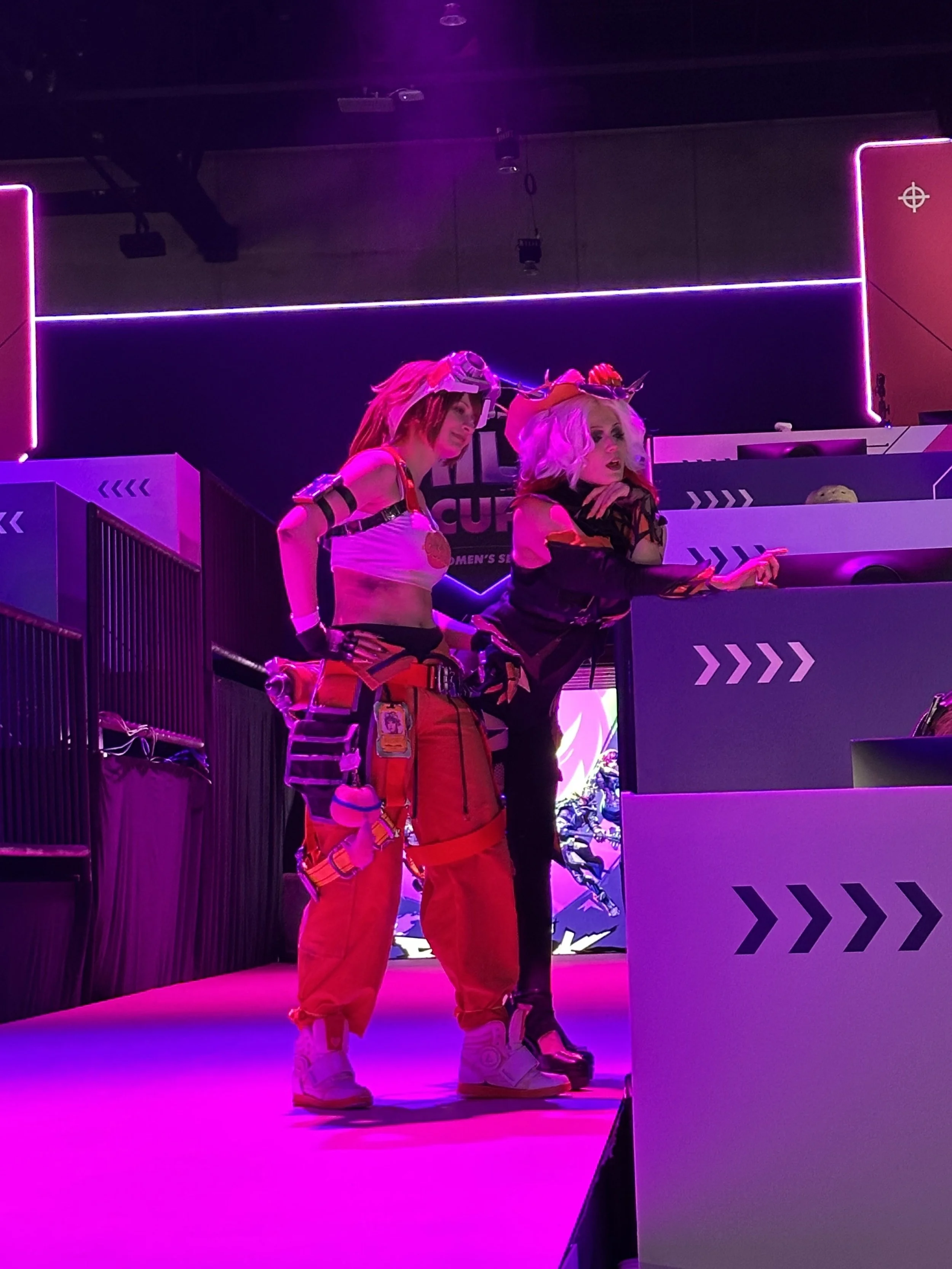 Two women dressed as characters from Overwatch esports event, with colorful costumes, wigs, and makeup, standing on stage with pink lighting, behind a barrier with directional arrows.