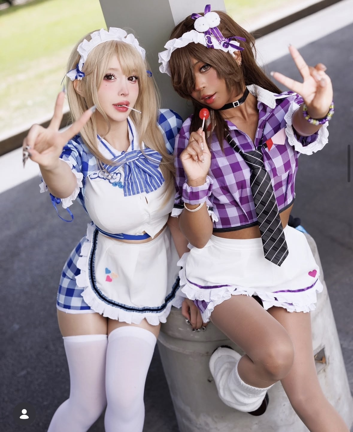 Two women dressed in cosplay nurse costumes with plaid patterns and ruffled aprons, posing and making peace signs while standing outside near a concrete structure.