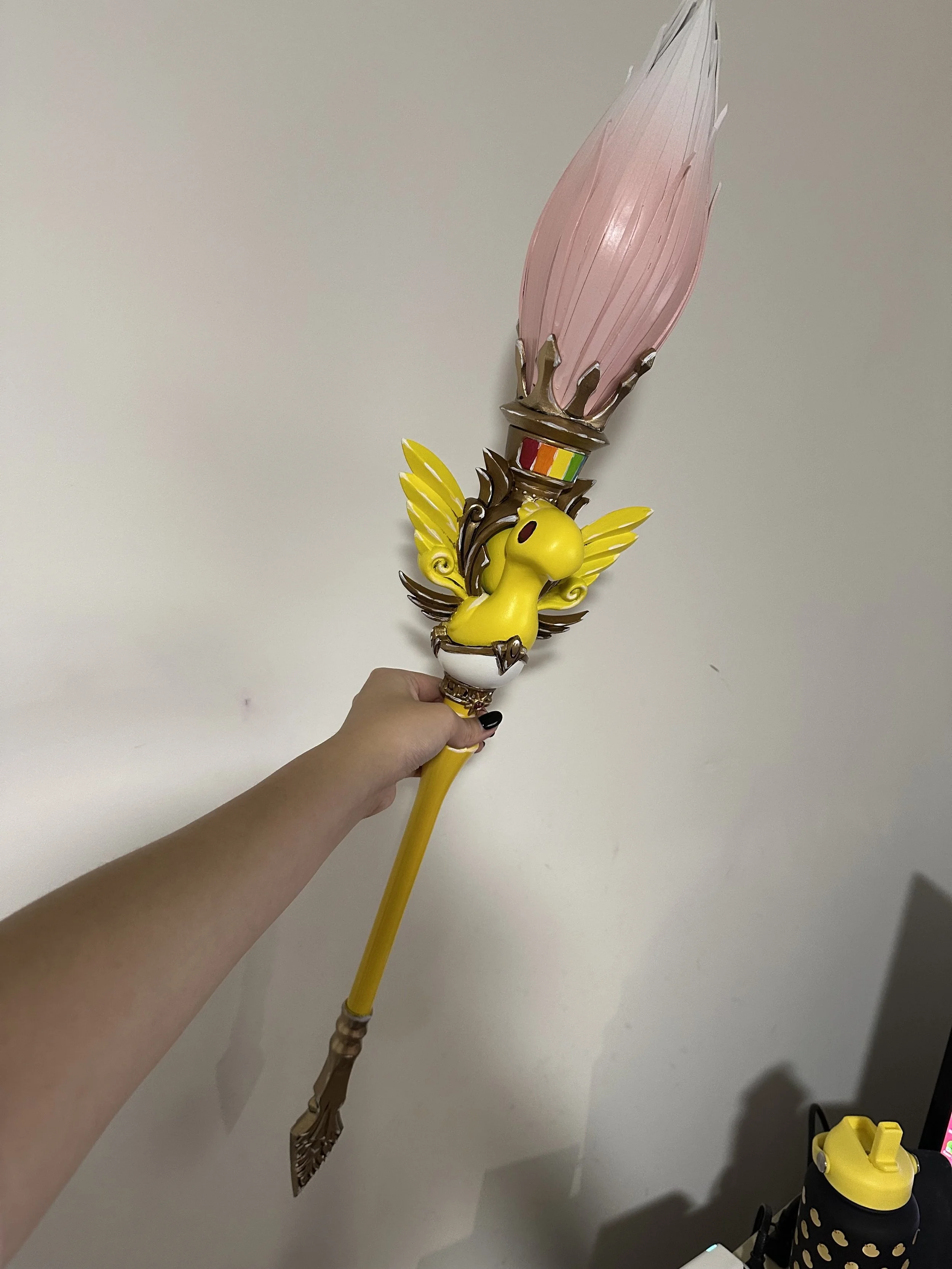 A hand holding a staff featuring a yellow character with wings and a pink flame at the top.