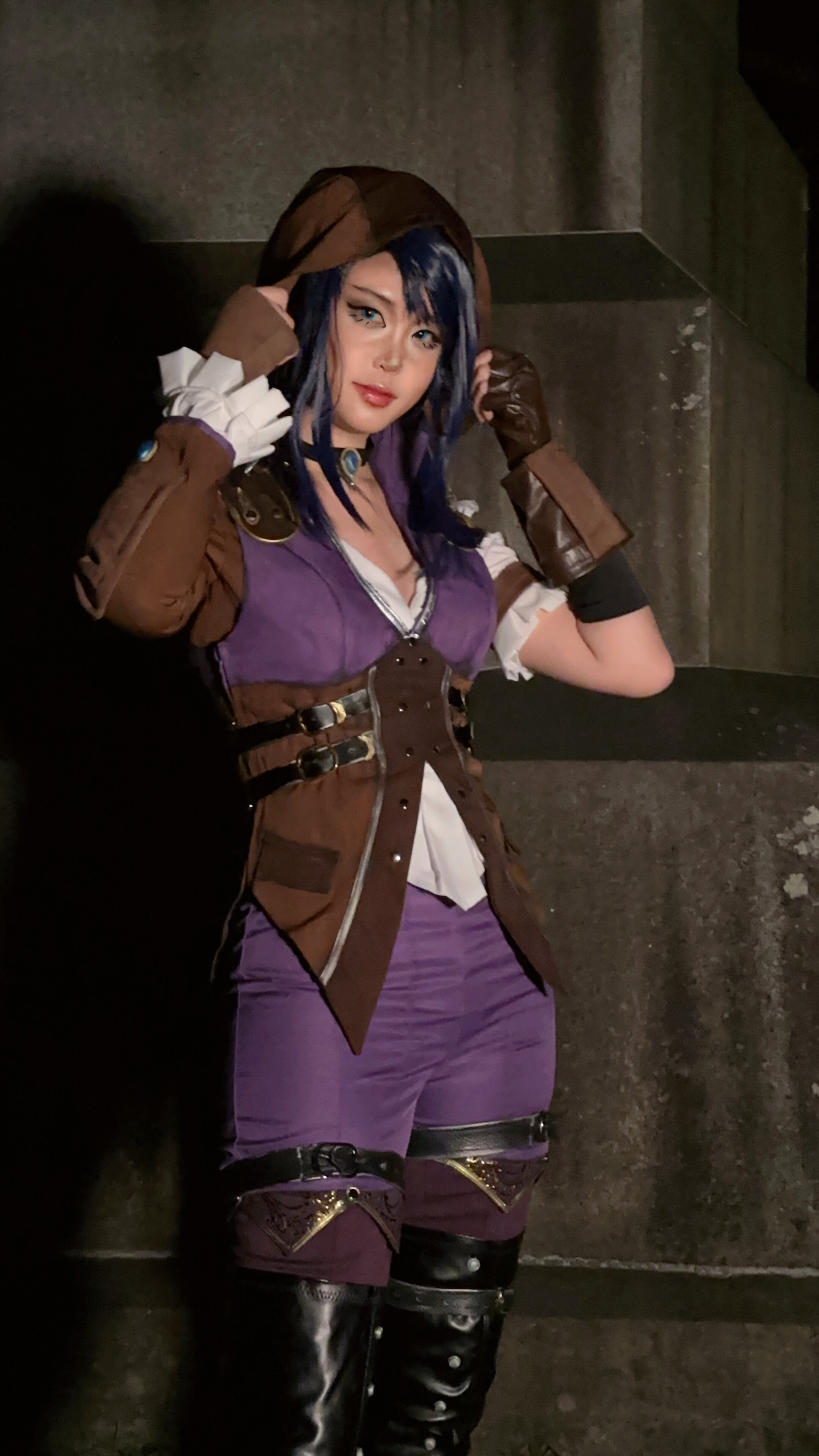 A woman dressed in a fantasy pirate or adventurer costume with purple pants, brown leather armor, gloves, and a hood, standing against a dark wall.