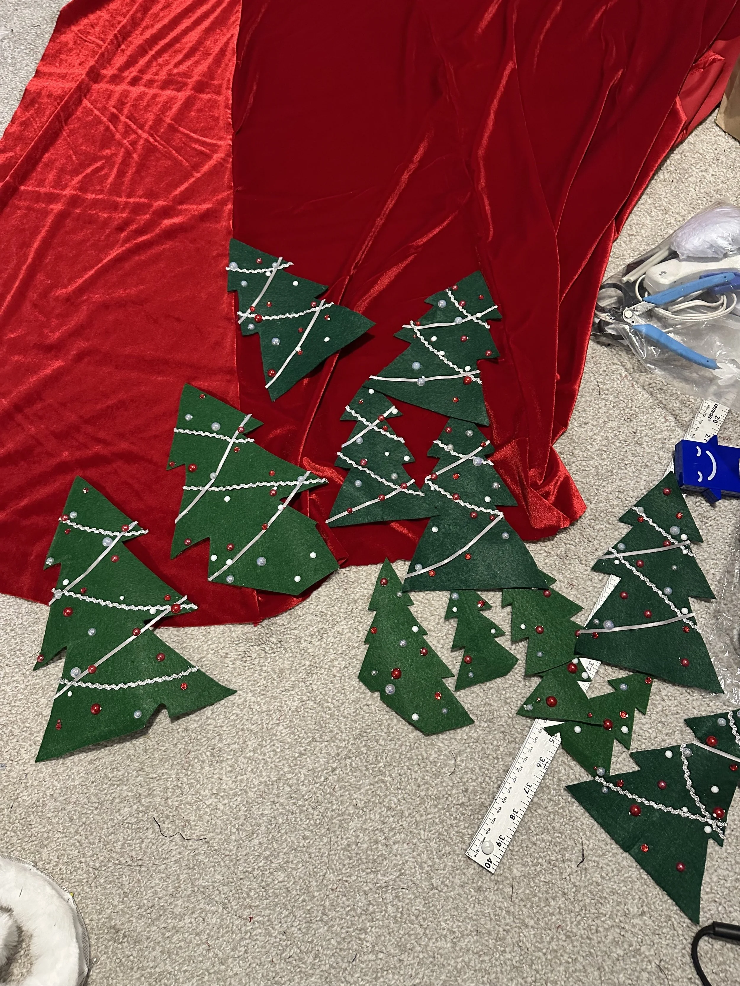 Several DIY Christmas tree decorations made of green felt in various sizes with red and silver beads and white string, placed on a beige carpet and red fabric background.