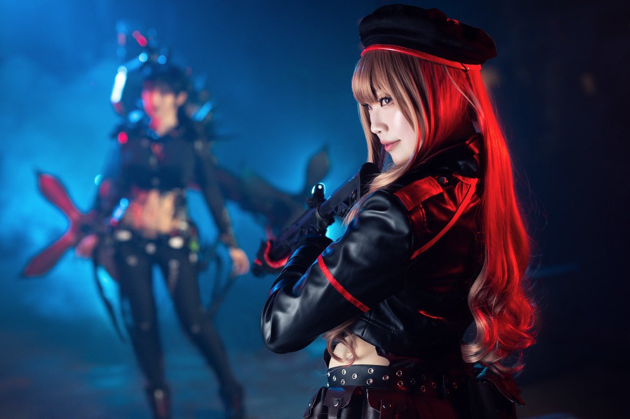 A woman with long, wavy hair and a confident expression, holding a gun in a dark, blue-lit setting with another woman dressed in elaborate costume in the background.