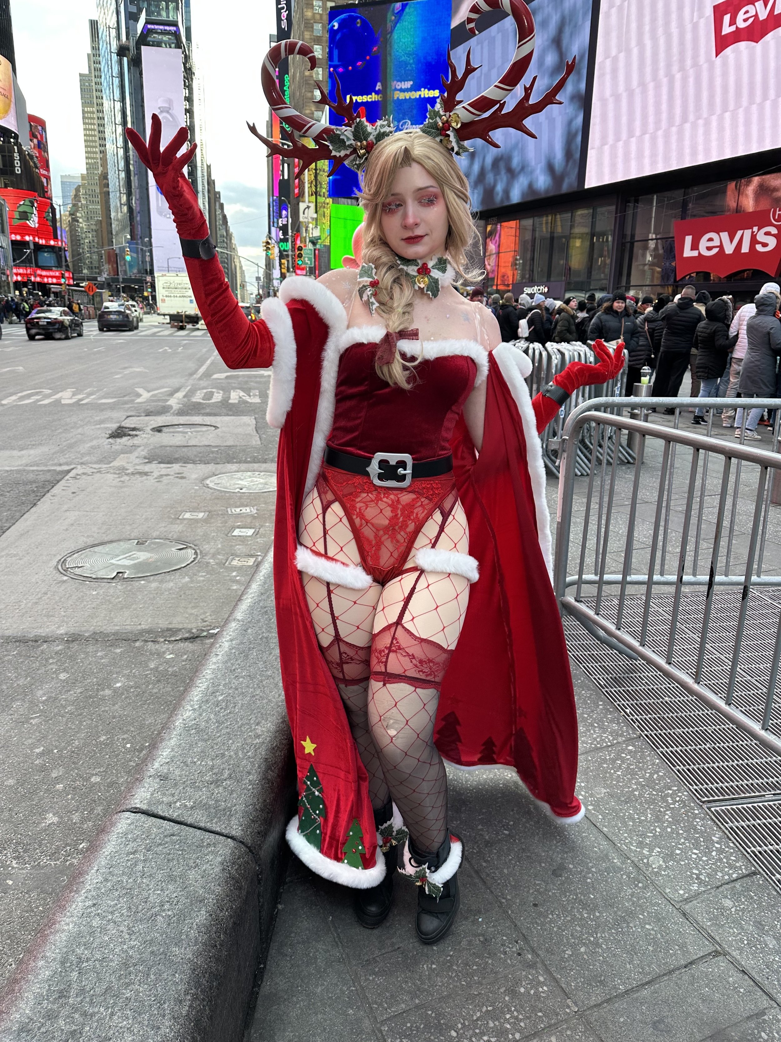 Woman dressed in festive Christmas attire, including a red cape with white trim, red fishnet stockings, red lace lingerie, and a headpiece with reindeer antlers, standing on a city sidewalk with Times Square in the background.