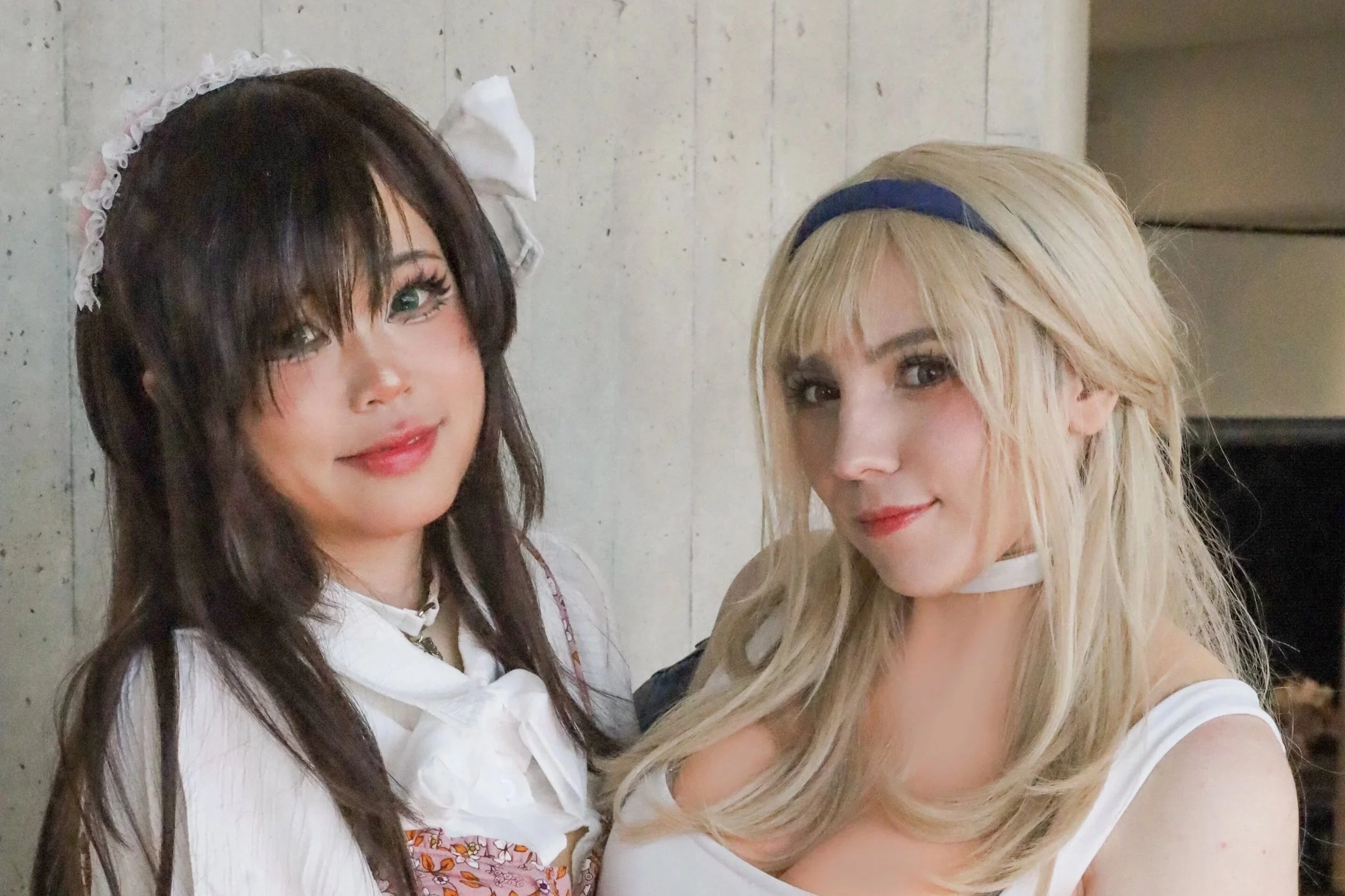Two women posing together indoors, one with dark hair and makeup resembling a doll, wearing a white blouse with a ruffled collar and a pink patterned vest, and the other with blonde hair and makeup, wearing a white top and a blue headband.