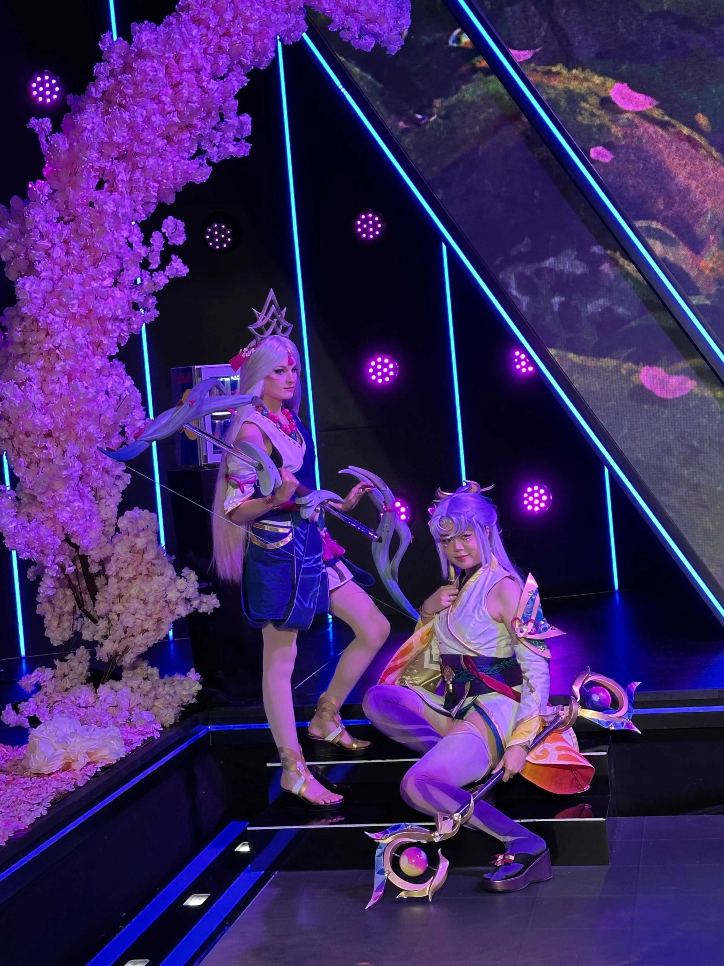 Two animated fantasy characters with elaborate costumes and weapons in a neon-lit display, highlighted by pink cherry blossom decorations.