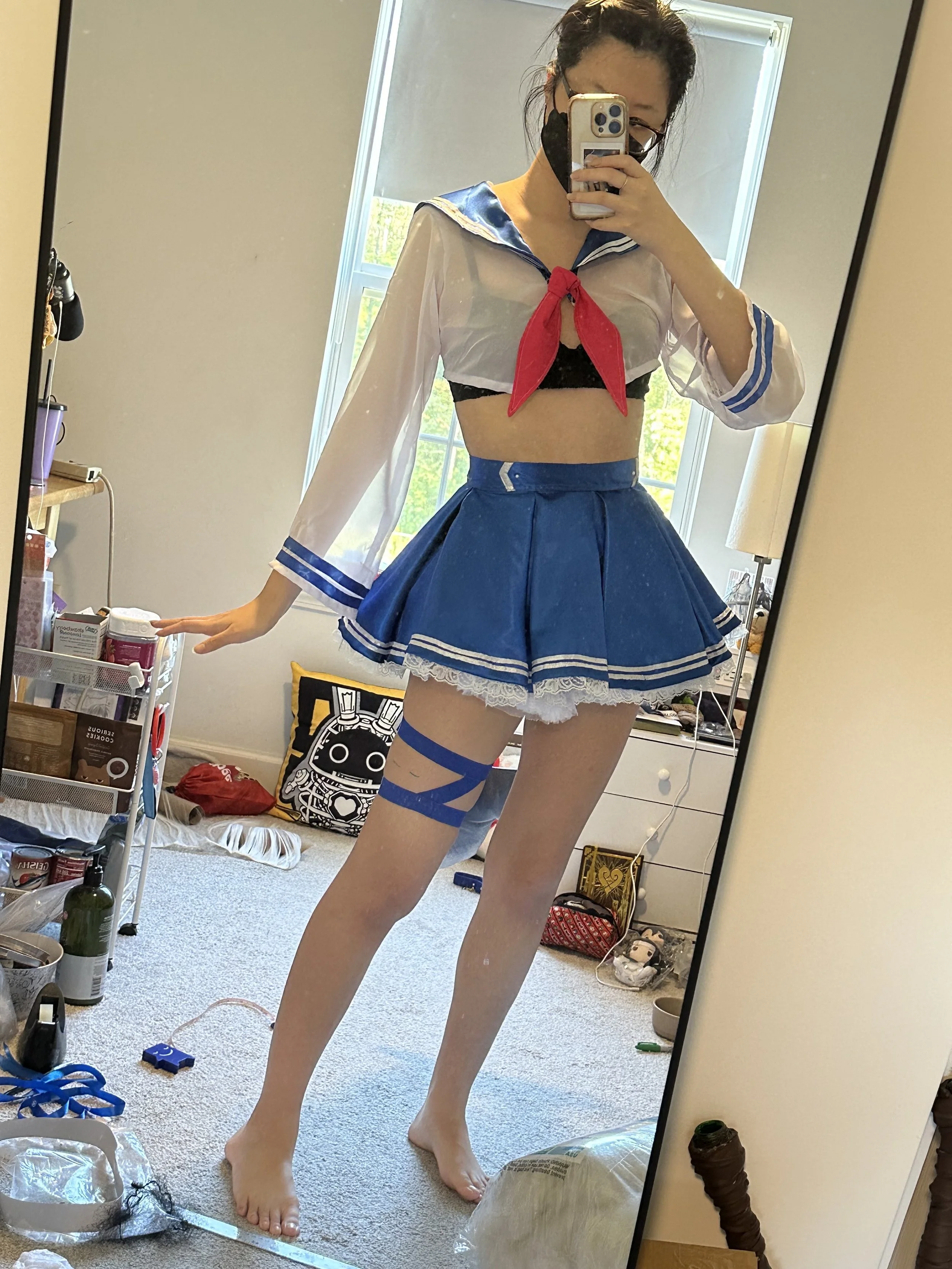 A person taking a mirror selfie is dressed in a revealing cosplay costume inspired by a sailor school uniform, with a blue skirt, white top with blue accents, black bra, and thigh-high stockings with blue straps. The person has dark hair, glasses, an