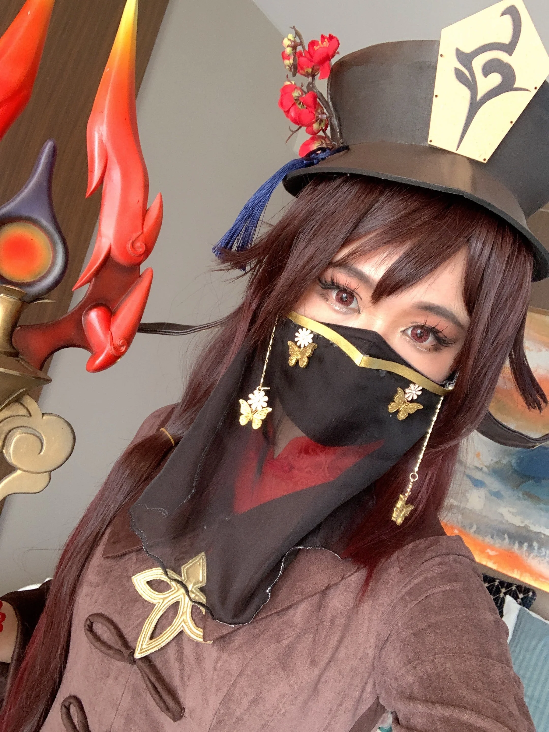 Person wearing a large black hat with red flowers and a gold emblem, a black face mask decorated with gold butterflies and white flowers, a brown costume with red details, standing in front of decorative artwork including a fiery red and black sculpture and a painting of waves.
