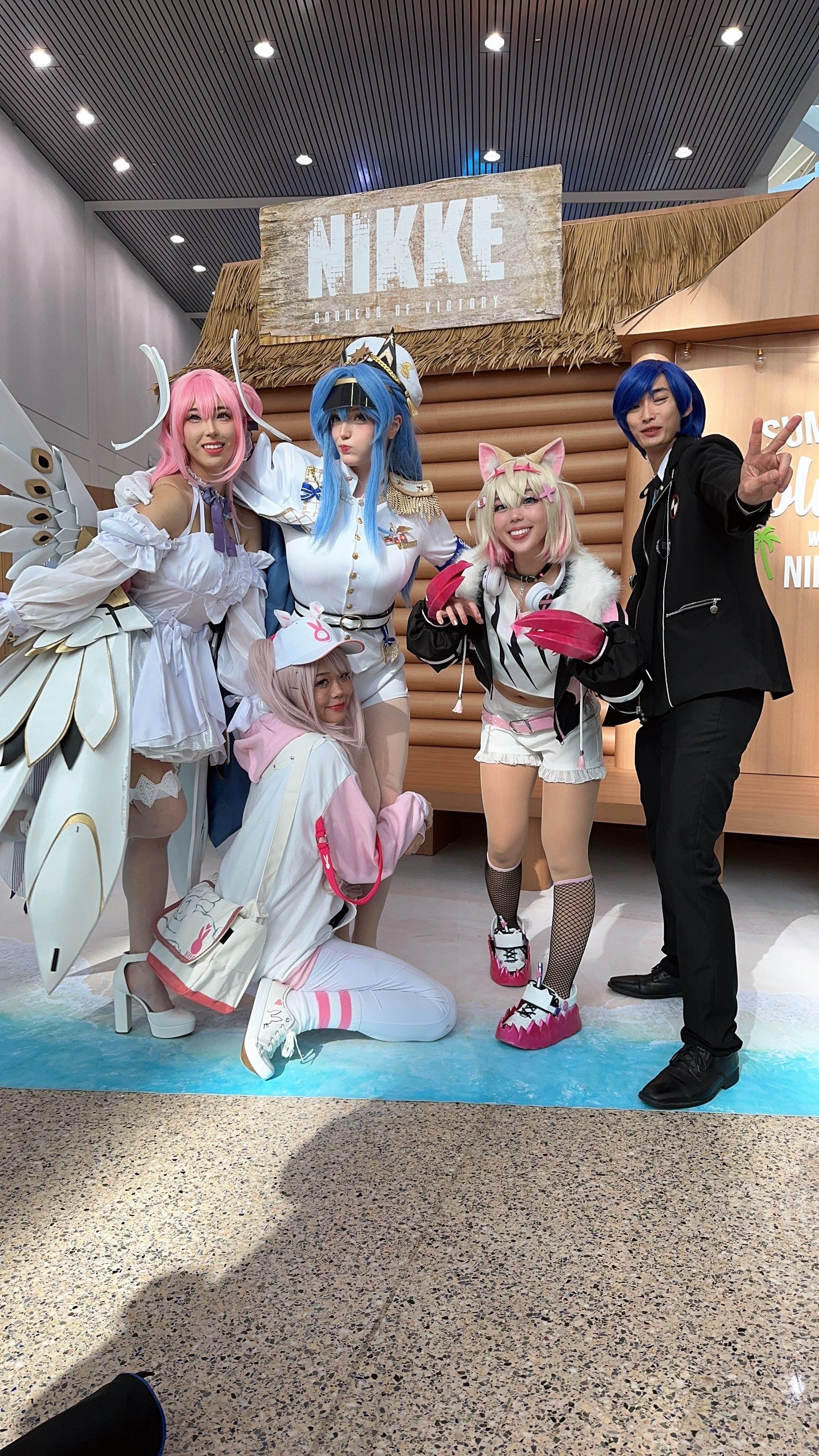 Group of five people in colorful cosplay costumes posing at an indoor event in front of a wooden sign that says "NIKKE" with smaller text "GATHER OF VICTORY" underneath.