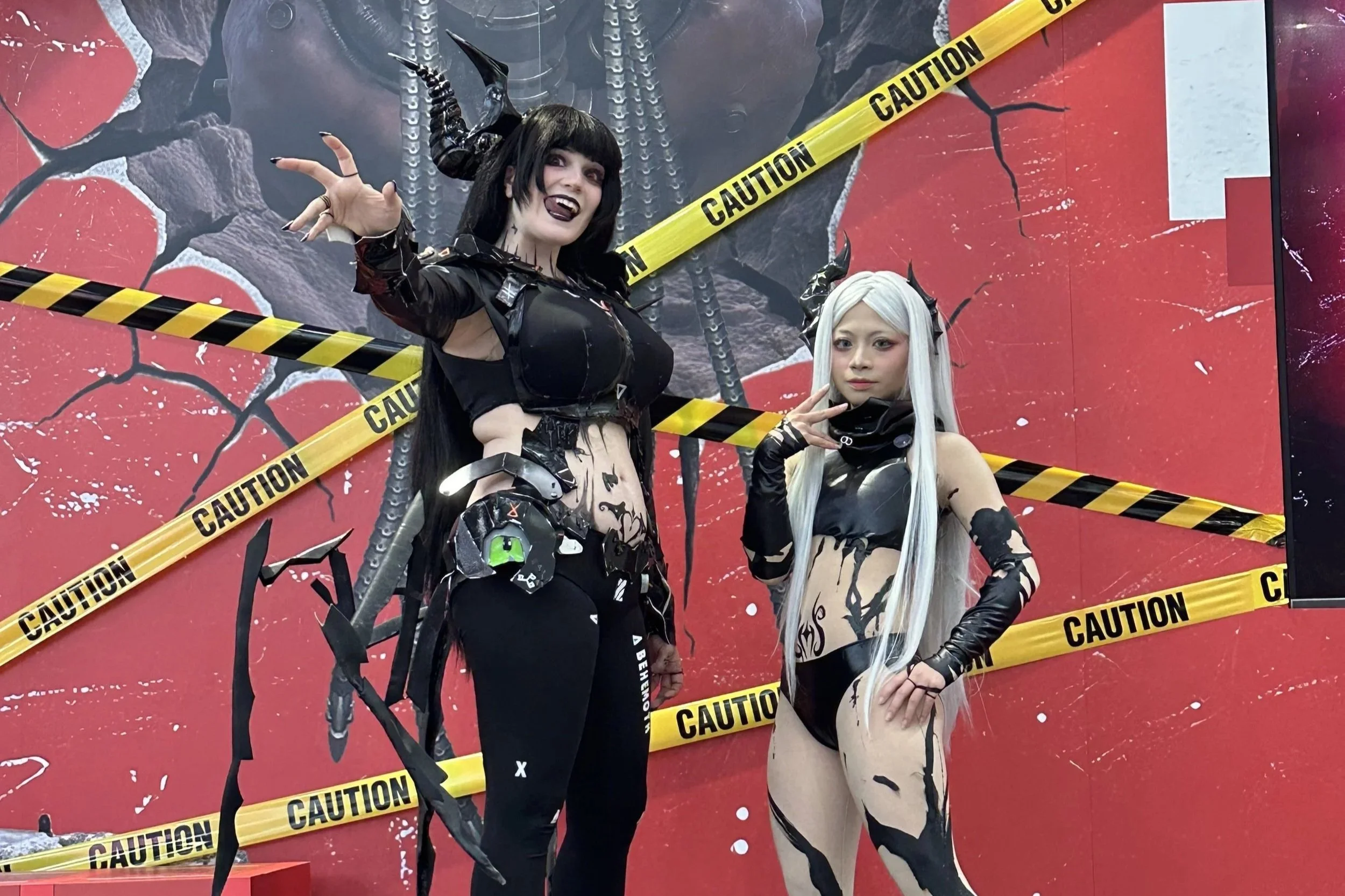 Two women in elaborate cosplay costumes with black and white themes, featuring horns and detailed makeup, standing in front of a red background with cracked patterns and yellow caution tape.