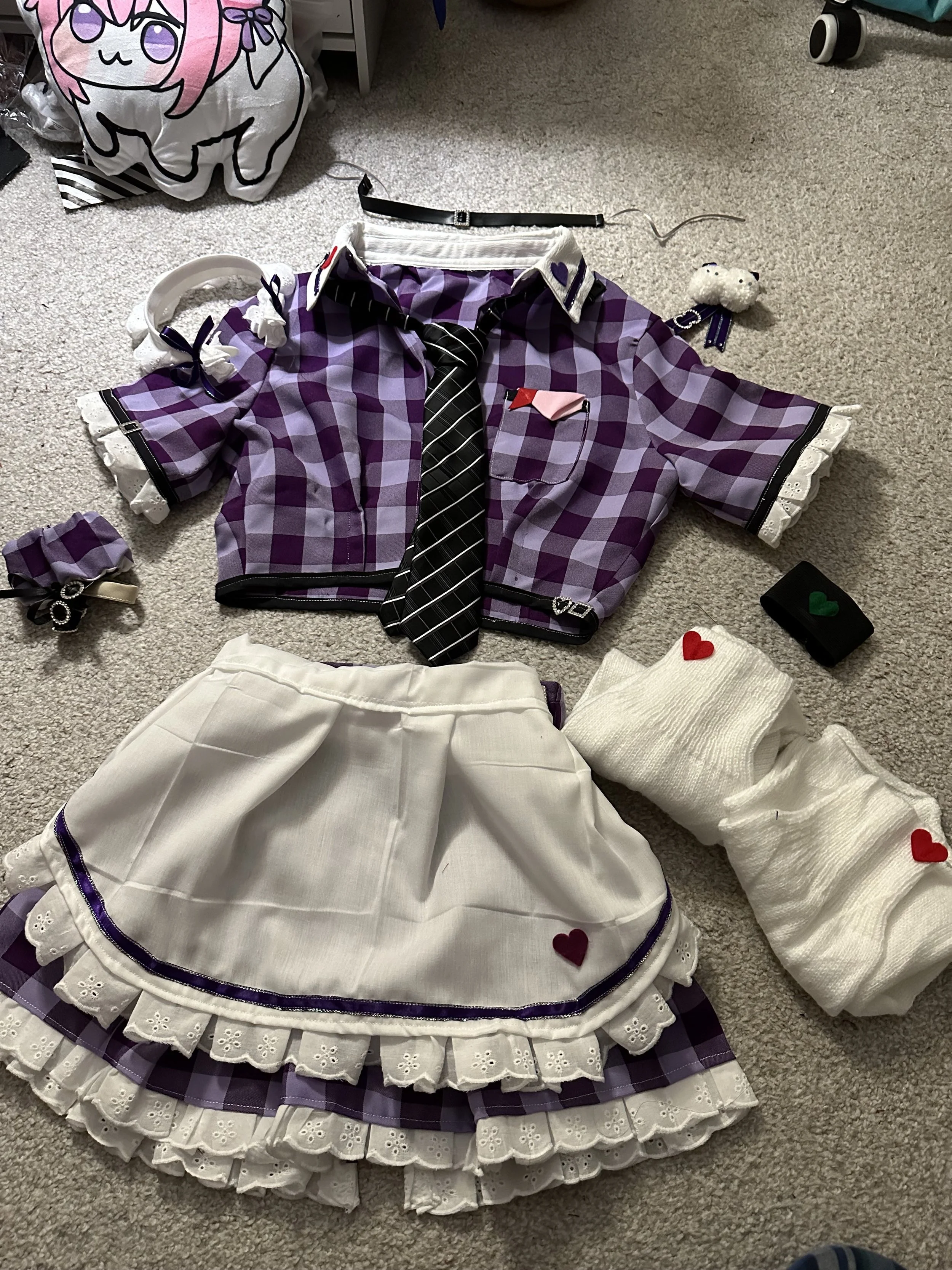 A collection of purple and white clothing items and accessories on a beige carpet, including a plaid cropped jacket, a plaid skirt with lace trim, a tie, a white sweater with hearts, a hairband, and various small accessories.