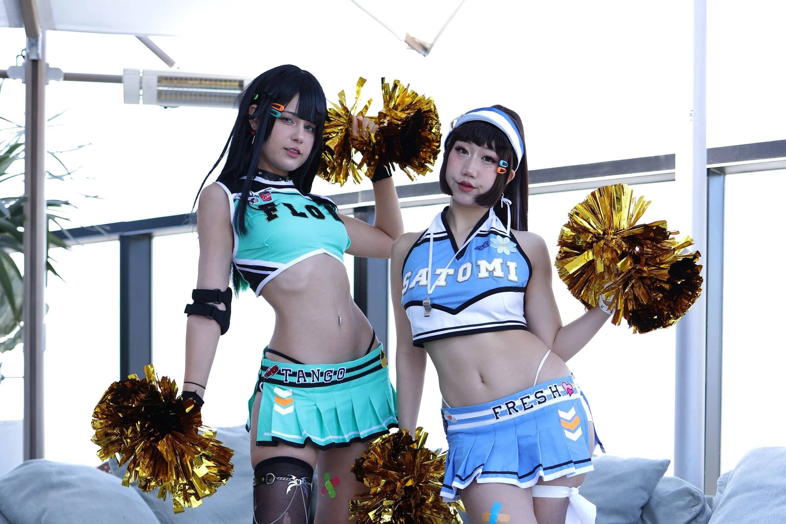 Two young women dressed as cheerleaders holding gold pom-poms, standing indoors with a large window behind them.