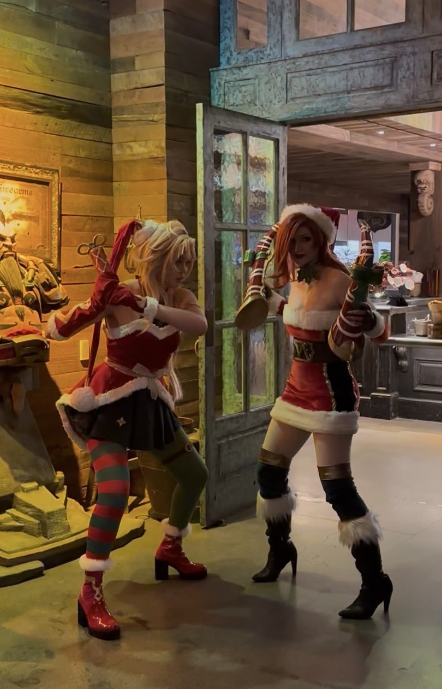 Two women dressed in Christmas-themed costumes, posing inside a rustic wooden room with a door open to a kitchen area, with holiday decorations and warm lighting.
