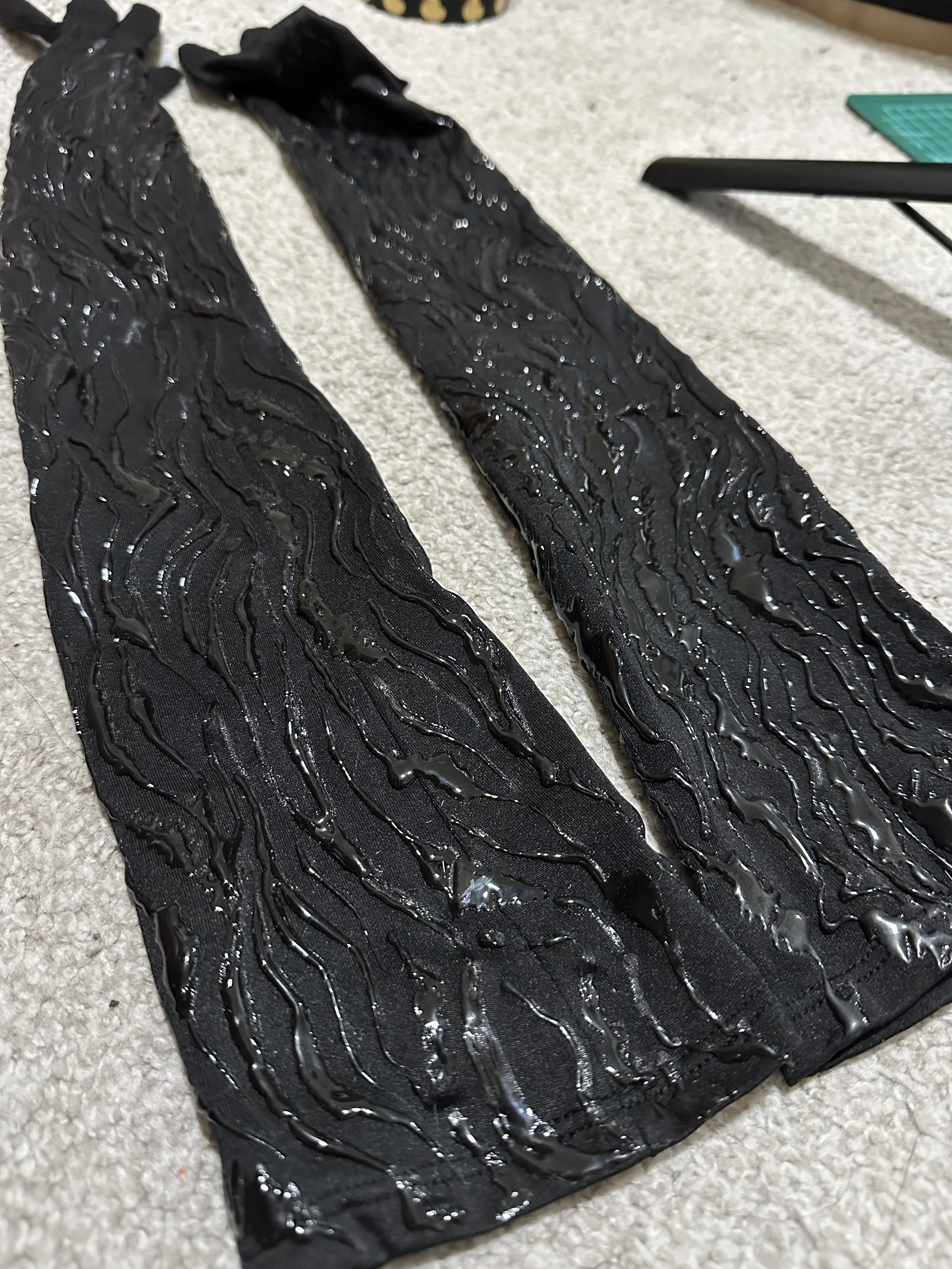 Two black fabric pieces with shiny, wavy, textured patterns laid out on a beige carpet.