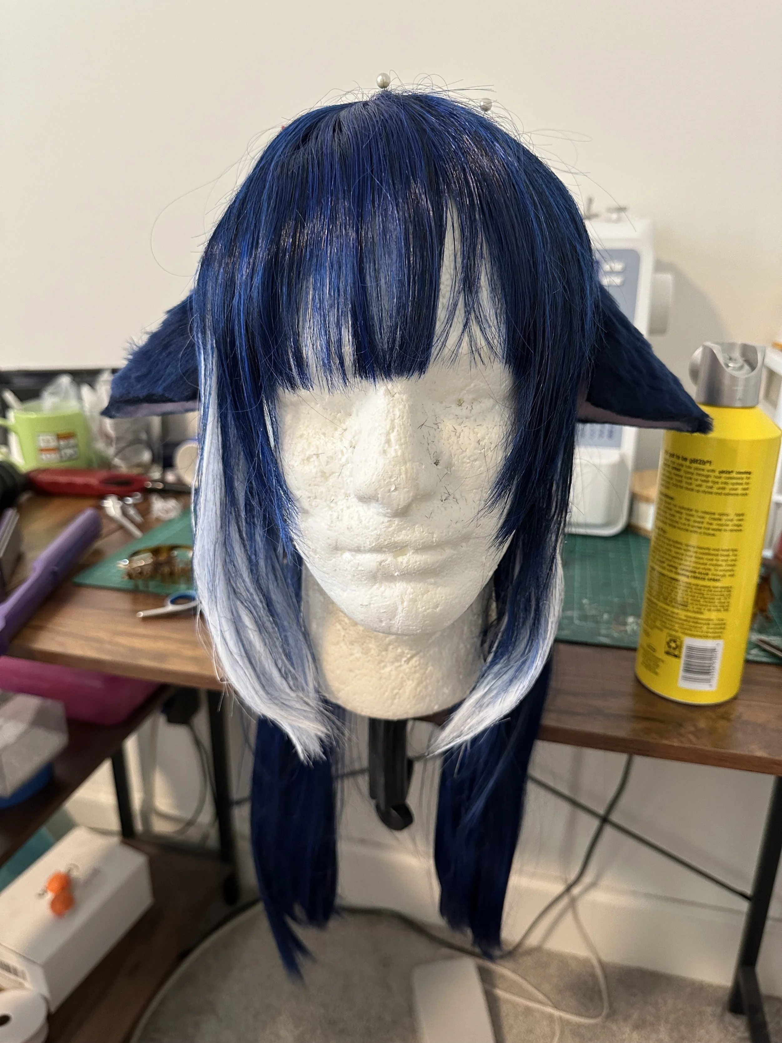 A mannequin head with dark blue and white styled wig, cat ears, and bangs, on a work table with tools and supplies in the background.