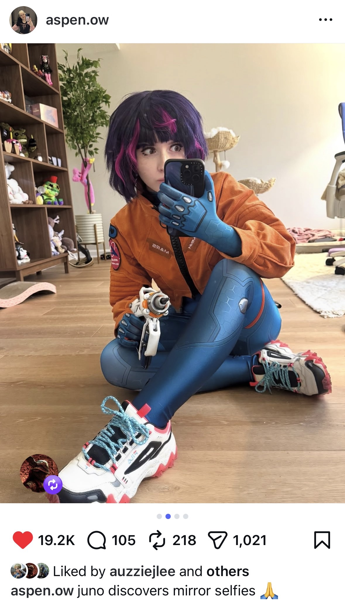 A person with purple hair and pink highlights taking a mirror selfie, wearing a brown jacket, blue tights, and sneakers, sitting on the floor of a room filled with shelves of toys, plants, and a cat tree.