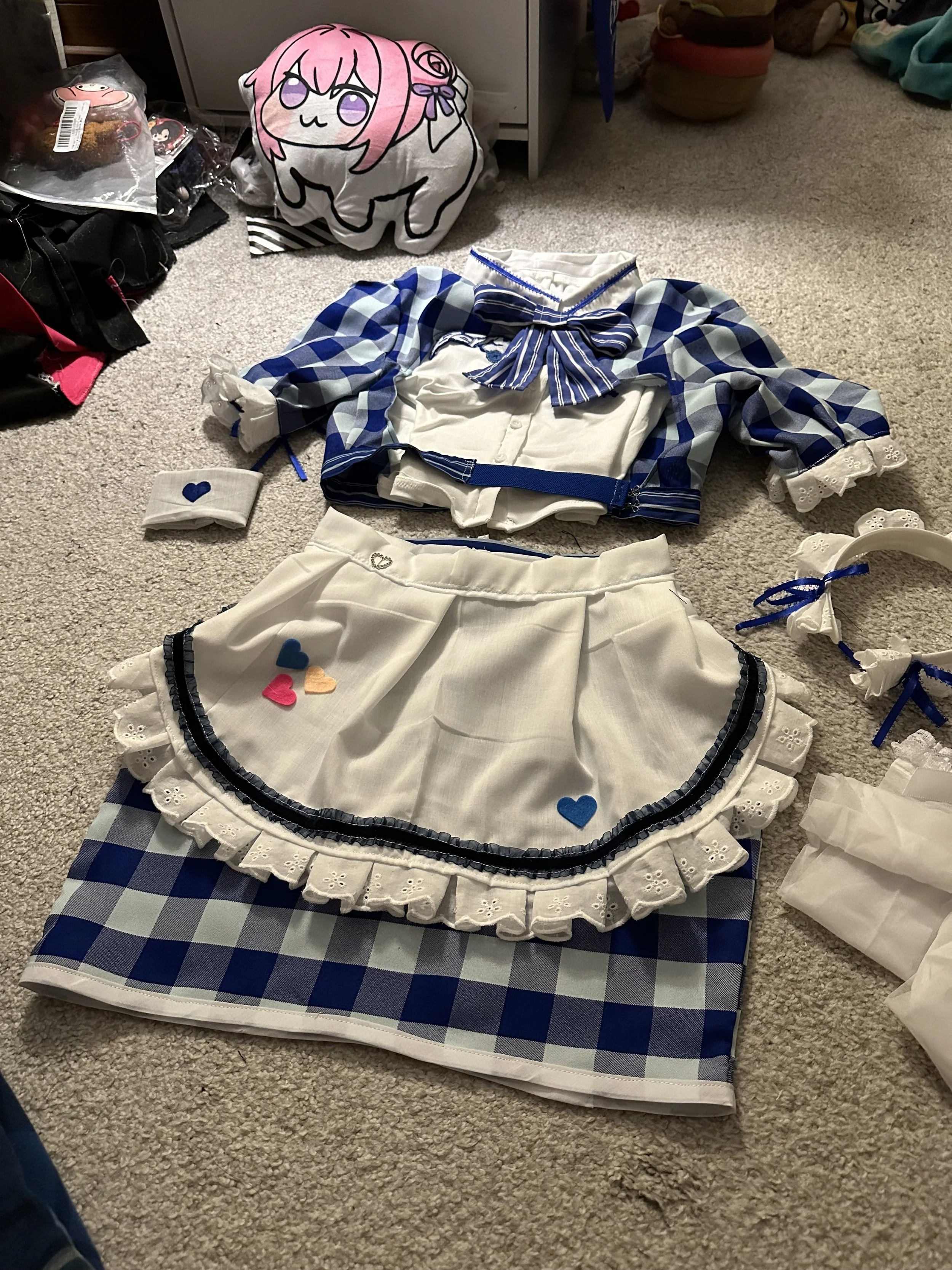 Two outfits laid out on a beige carpeted floor, both featuring blue and white checkered patterns and lace trimmings. The upper outfit is a jacket with a blue checkered bow, and the lower outfit is a skirt with small multicolored heart patches. Surrou