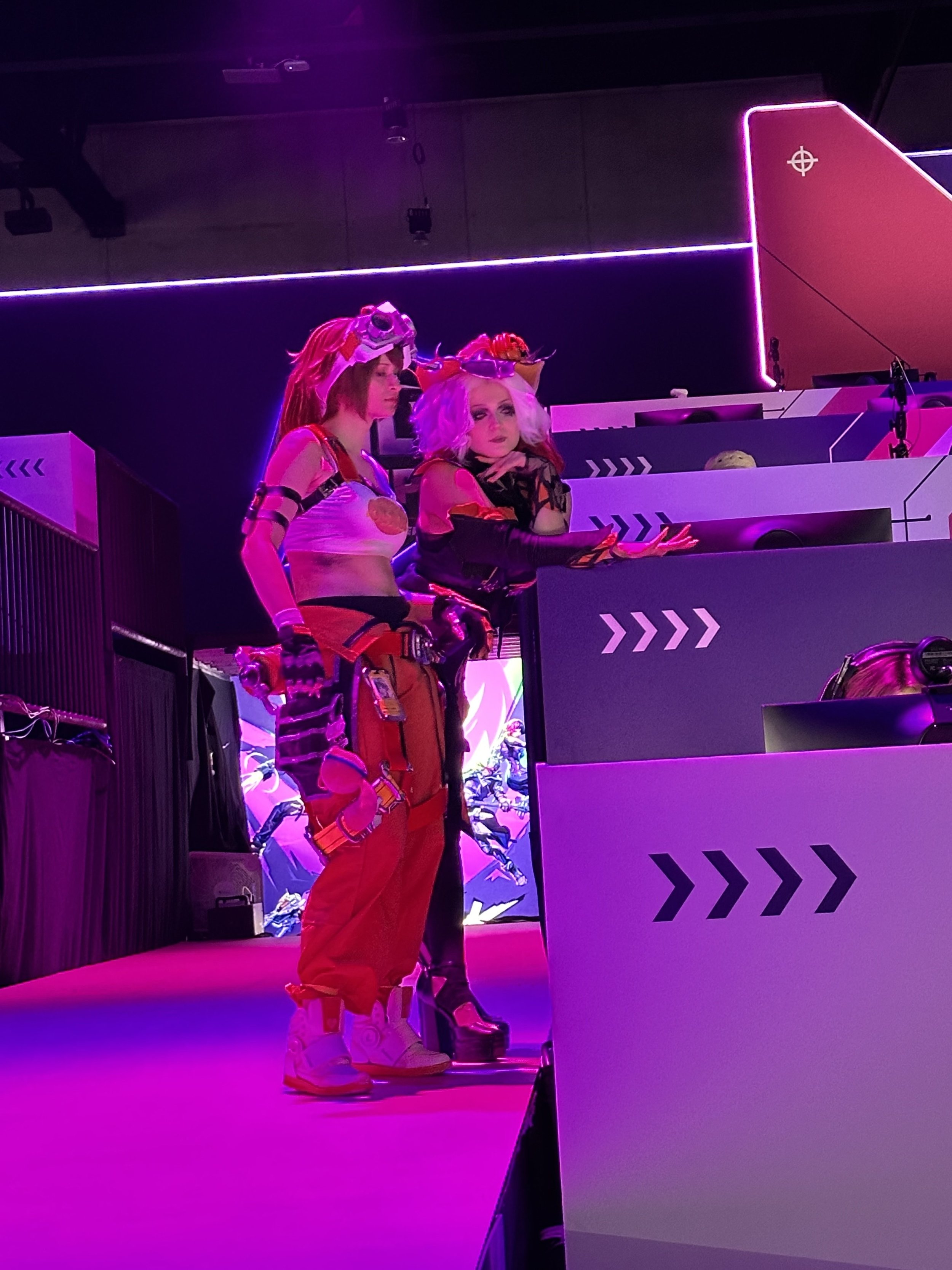 Two women in colorful costumes, one with bright pink and red hair wearing goggles, and the other with white hair wearing a crown, standing behind a row of gaming stations with purple and pink lighting.