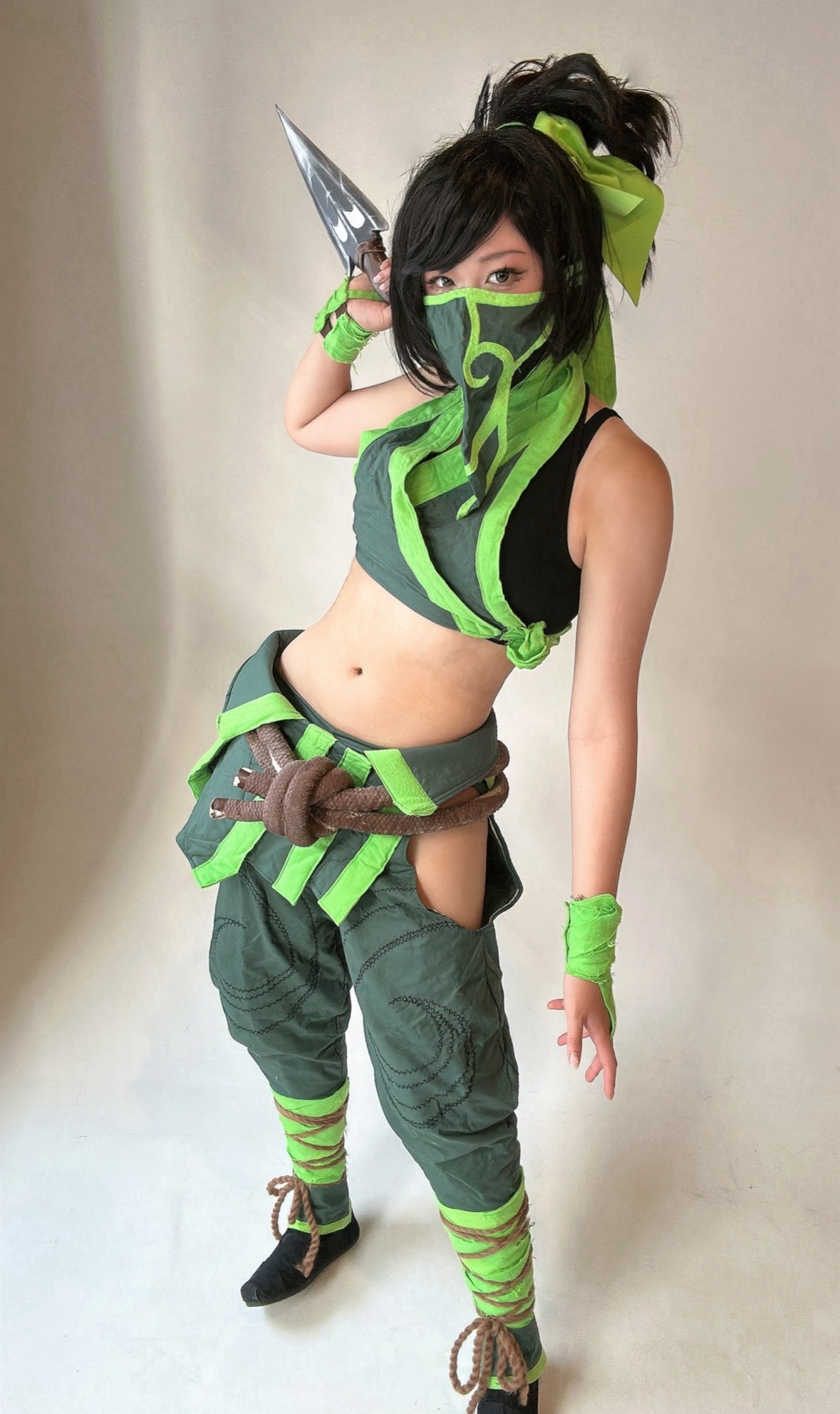 Person dressed in a green and black costume with a mask, holding a dagger, posing in a fighting stance.